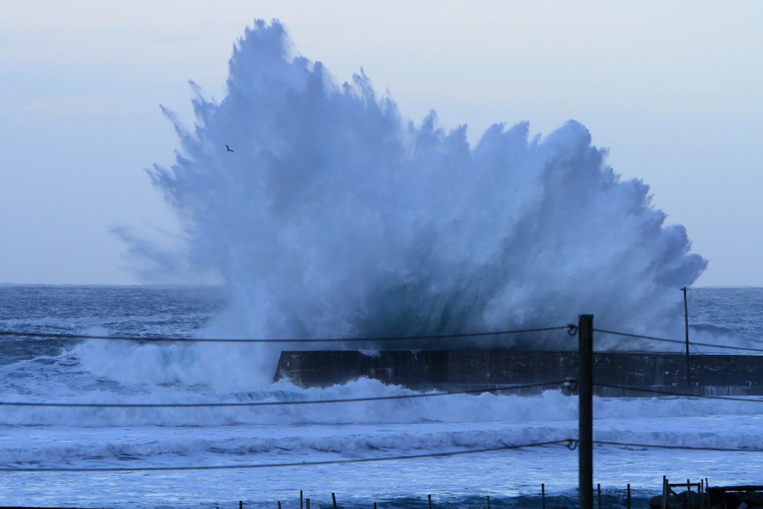 Large ocean wave crashing against a structure with water spray, under cloudy sky.