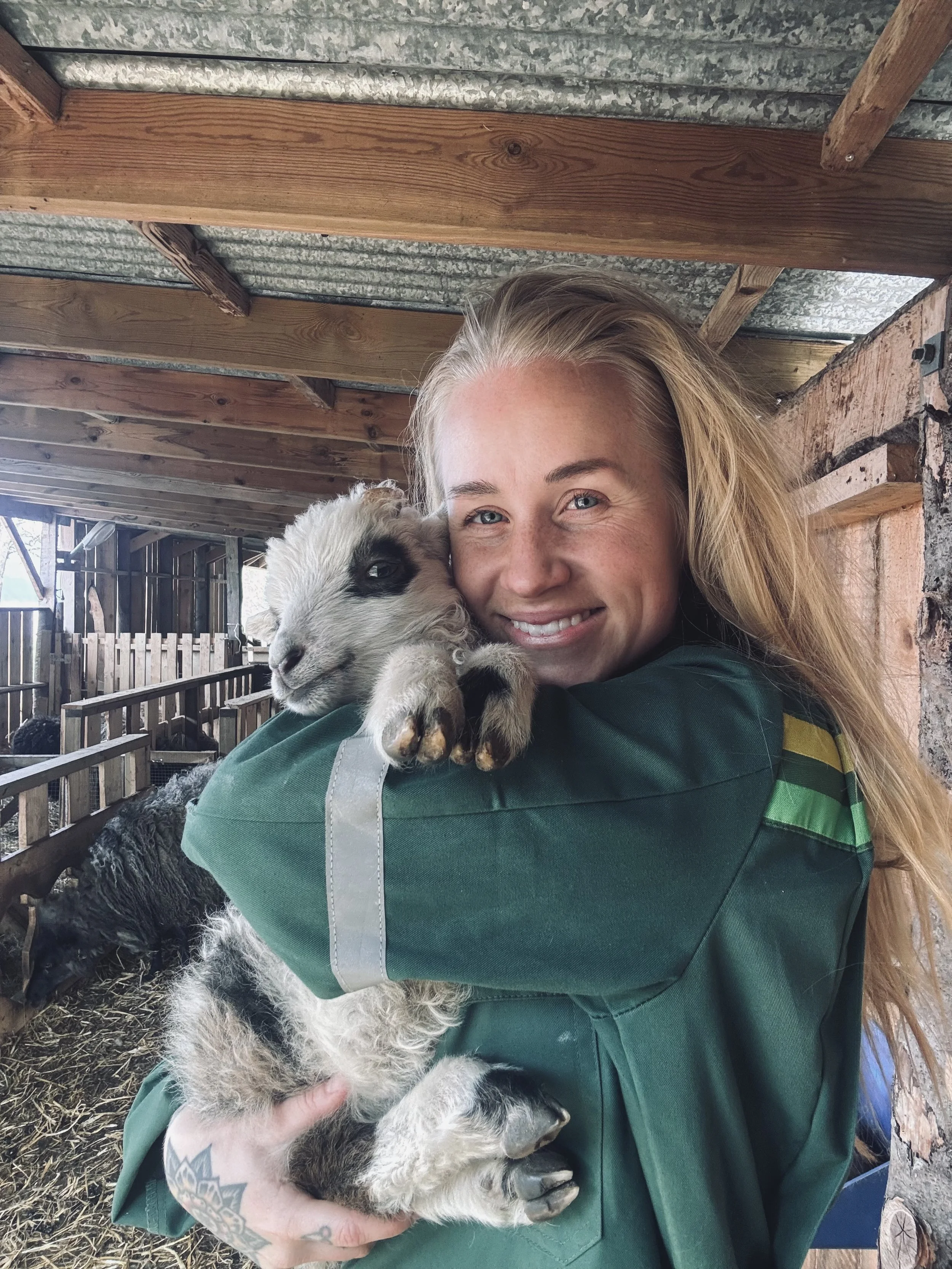 A woman with blonde hair smiling while holding a baby goat with black and white fur, inside a rustic wooden barn.