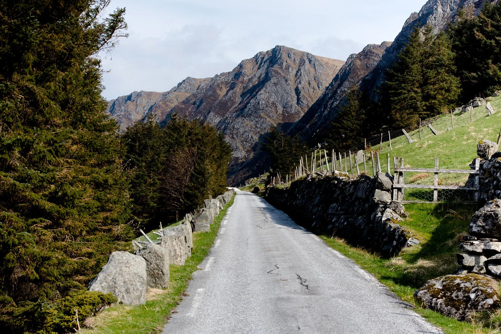 A narrow country road running through a mountainous landscape with green grassy slopes, stone fences, and dense trees on both sides, leading towards rocky mountain peaks in the distance under a partly cloudy sky.