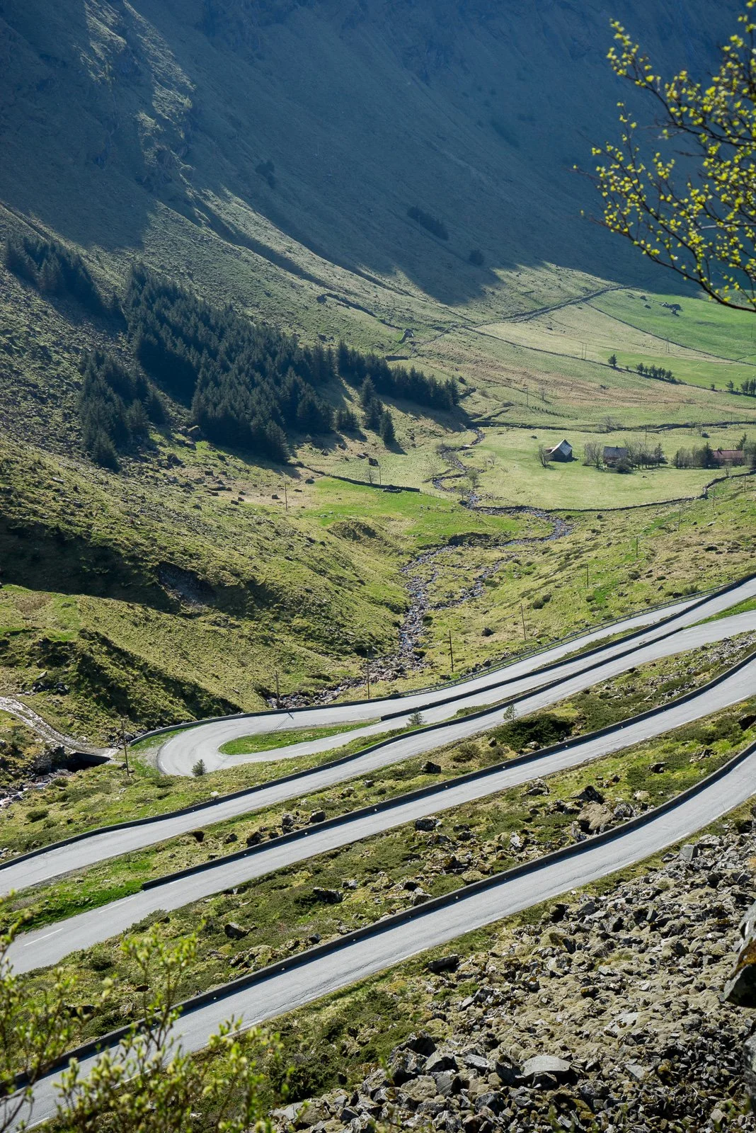 Winding mountain road with sharp curves, green grassy hills, trees, small houses, and a stream flowing through a valley in a mountainous landscape.