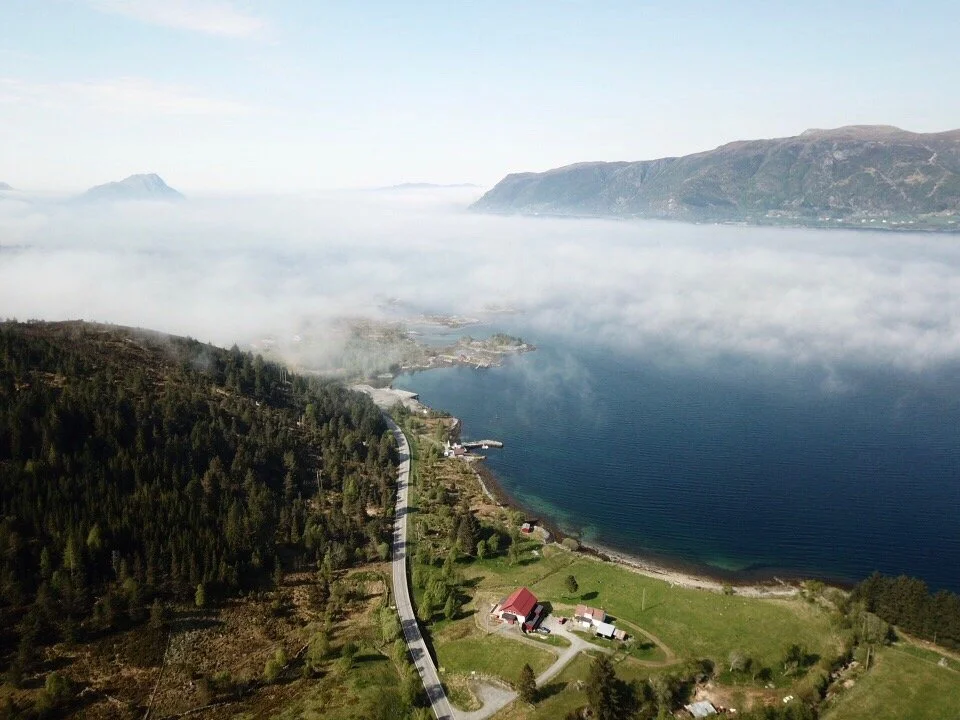 Aerial view of a scenic coastal landscape with mountains, a body of water, a winding road, and a few houses surrounded by greenery.