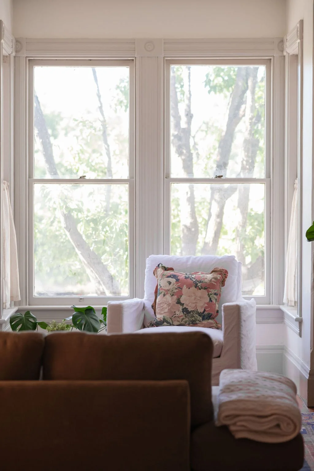 Sunlit room with large windows and white trim, white armchair with a floral pillow, green houseplants on the windowsill, and a folded blanket on a brown sofa.