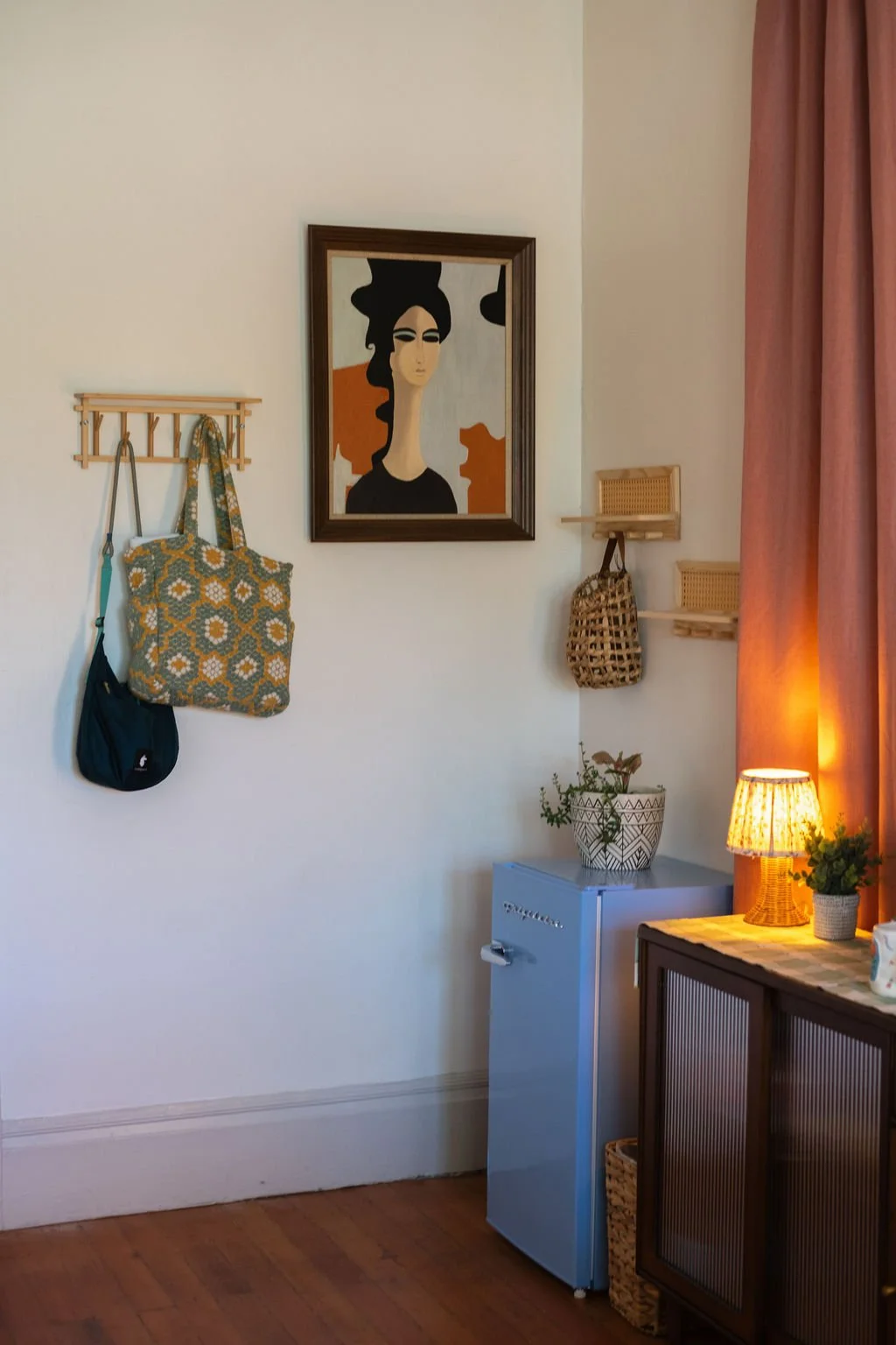 A corner of a living room with a small blue refrigerator, a framed portrait of a woman with dark hair on the wall, wall hooks with bags hanging, a small wooden shelf, a potted plant, a lamp on a vintage cabinet, and a pink curtain.