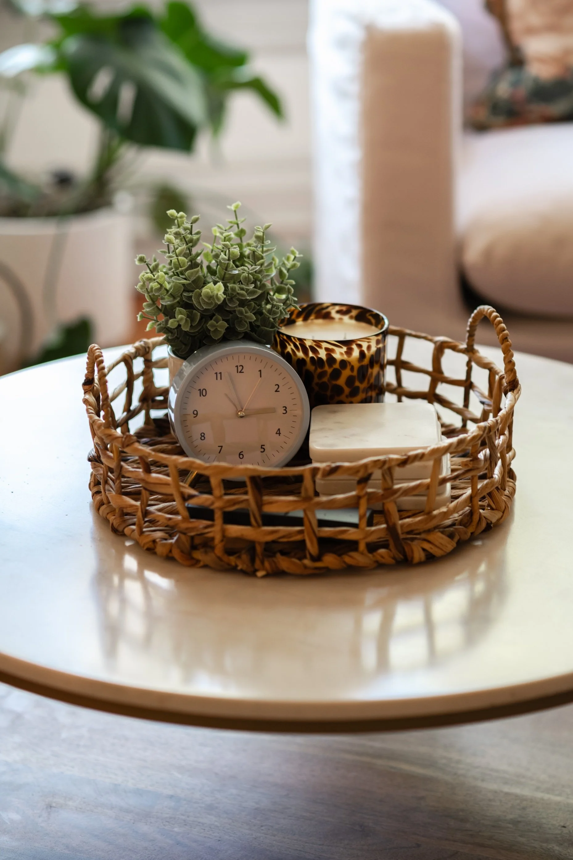 A wicker tray on a round table holds a small potted plant, a leopard print candle holder with a white candle, a white clock, and a small box, with a blurred background of a plant and furniture.