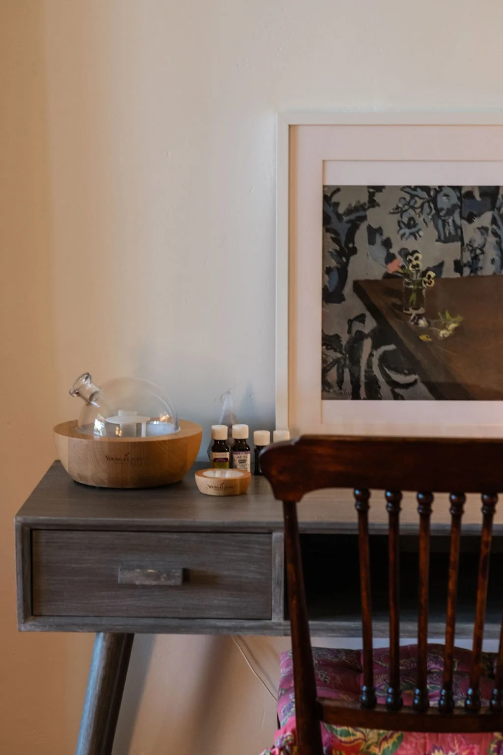 A wooden table with small bottles of essential oils, a glass bowl with glassware, and a white framed mirror reflecting a patterned rug and a wooden table with a flower arrangement.
