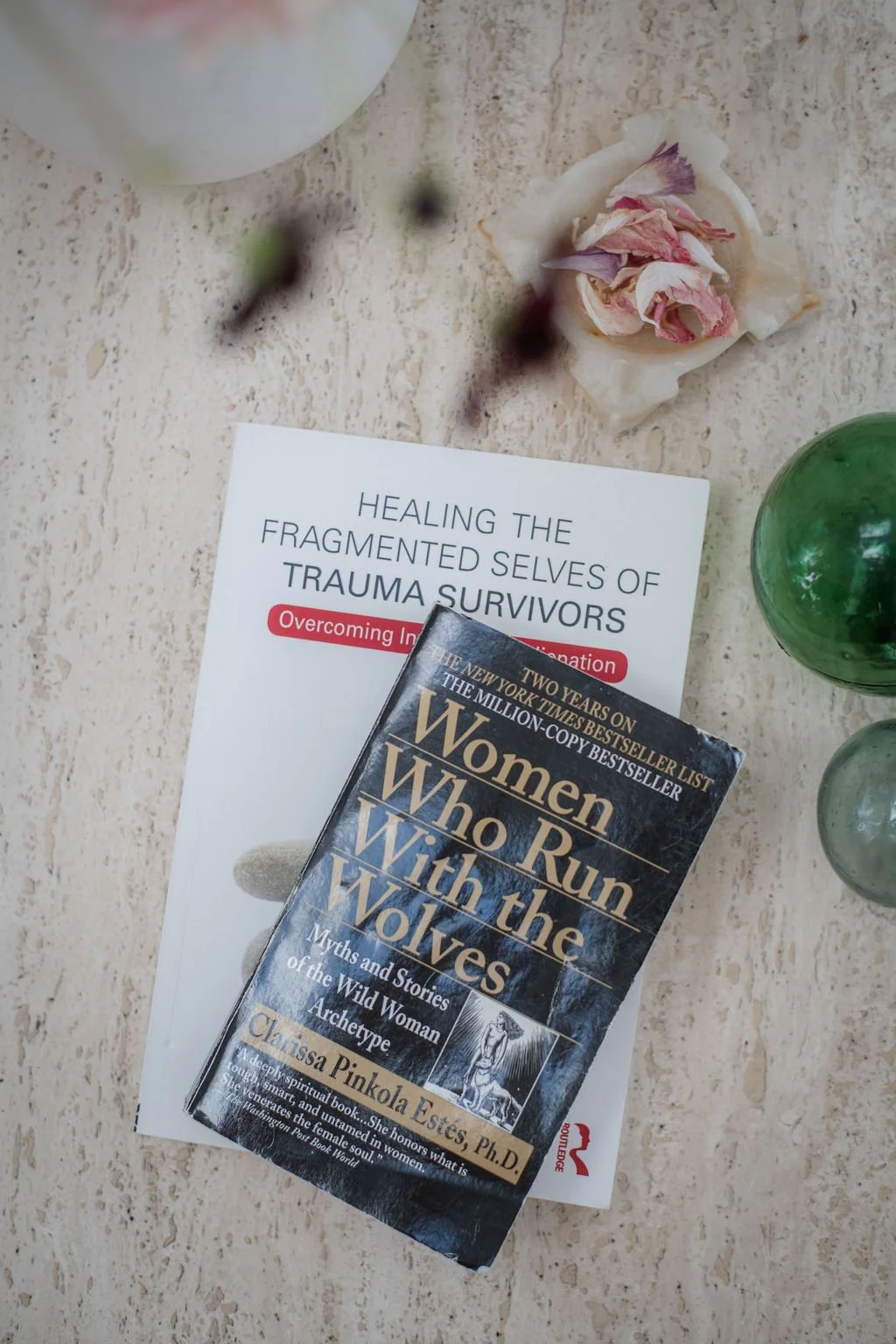 Books on trauma recovery and women in history, placed on a light-colored surface with a dried flower and green marbles nearby.