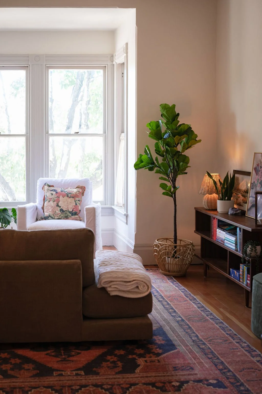 Cozy living room corner with a white armchair, floral pillow, tall green plant in a wicker basket, room beside large windows, and a wooden bookshelf with framed photos, a lamp, and books, illuminated by soft lighting.