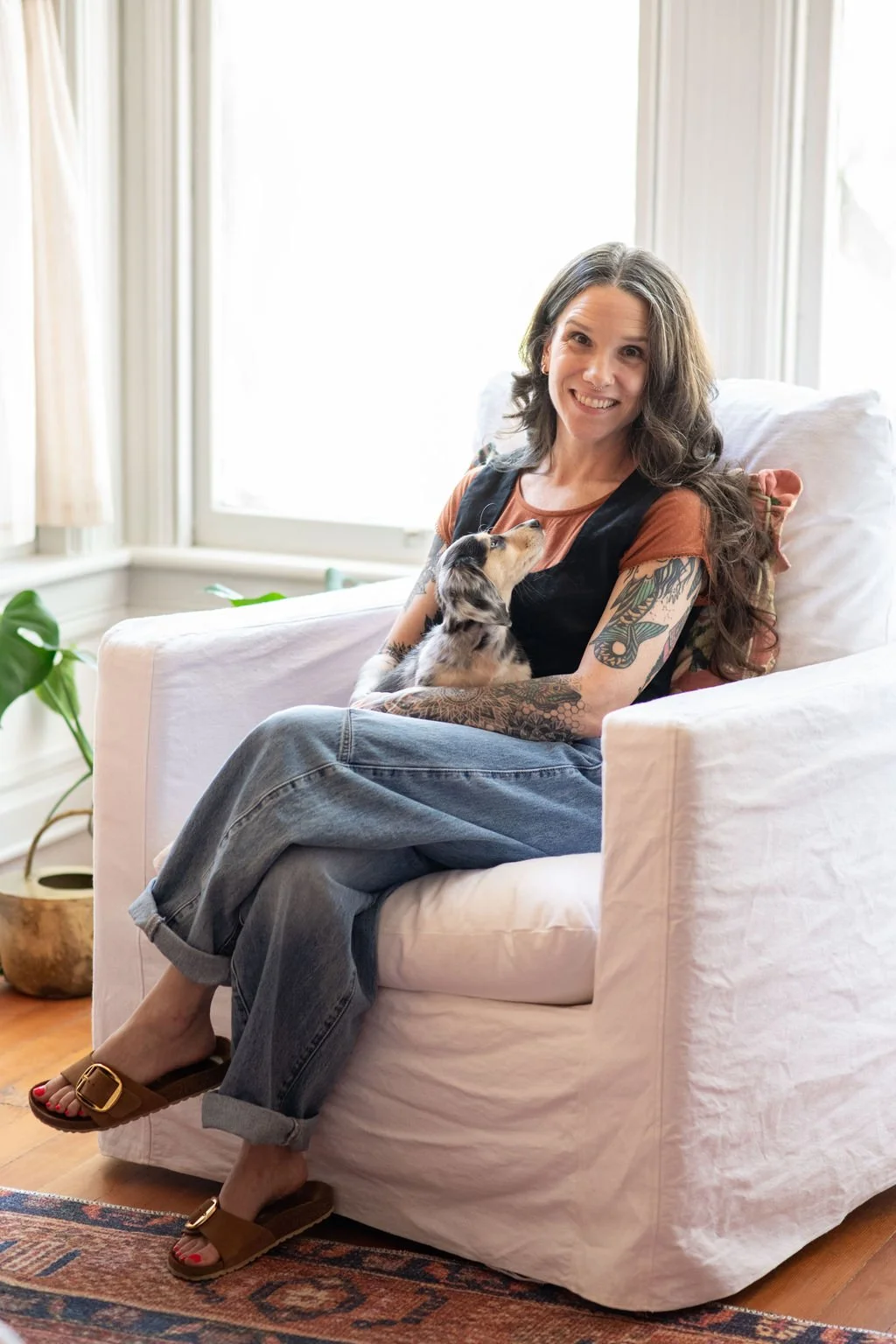 A woman with long wavy hair, tattoos on her arms, and a big smile sitting on a white couch with a small puppy. The room has large windows with white curtains, and there is a green plant nearby.