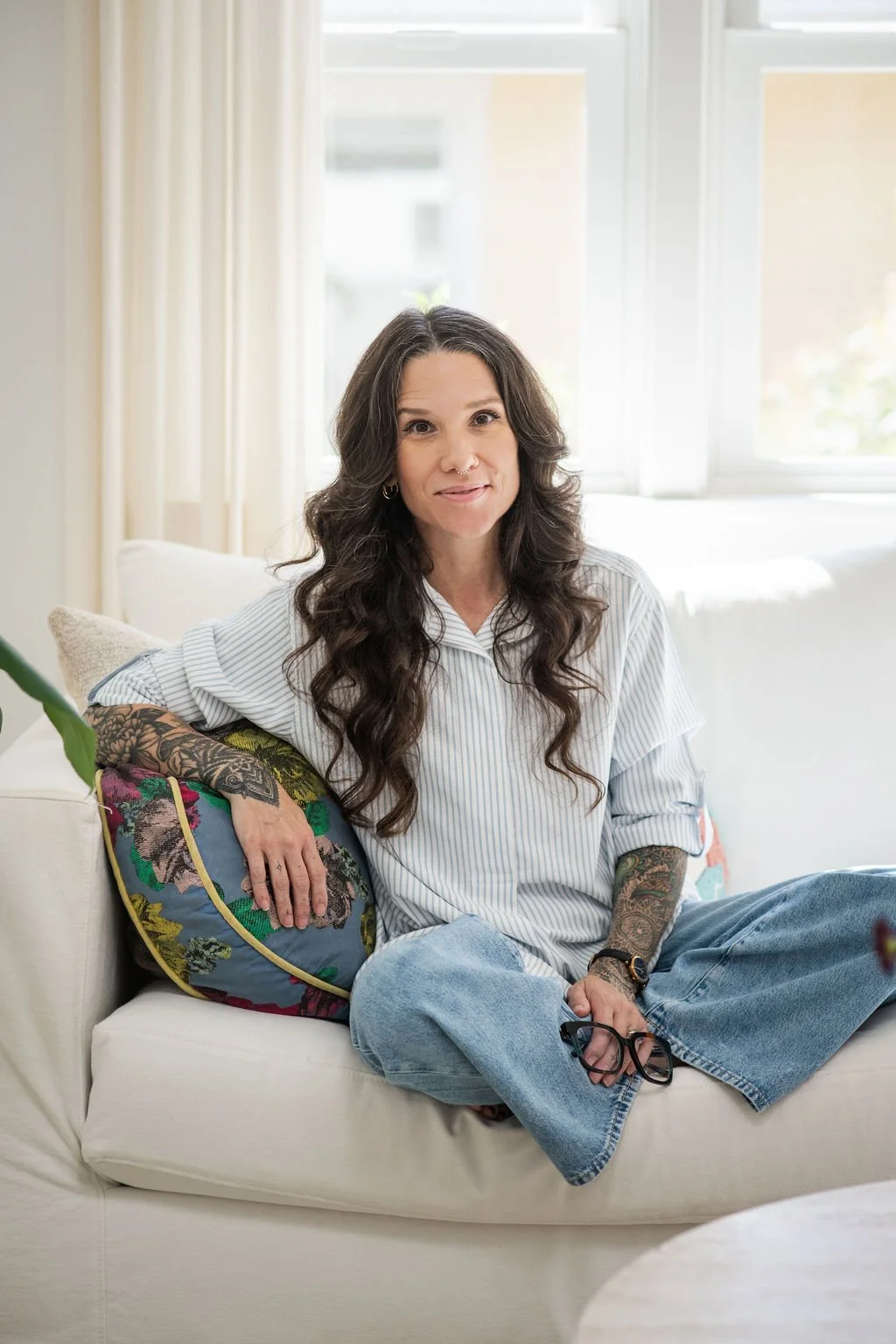 A woman with long, dark, curly hair and tattoos on her arms, wearing a striped button-up shirt and blue jeans, sitting on a white sofa with a colorful cushion, in a bright room with large windows.