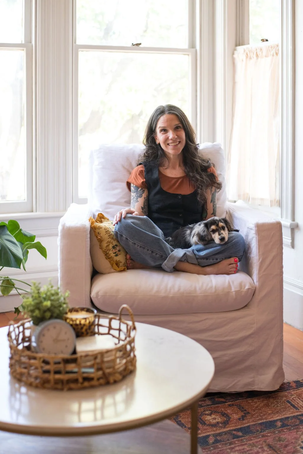 A woman with dark, wavy hair and tattoos on her arms sitting cross-legged on a beige armchair with a small dog resting on her lap, smiling in a bright living room with large windows, curtains, a plant, and a woven tray with a clock and a candle on a white coffee table.