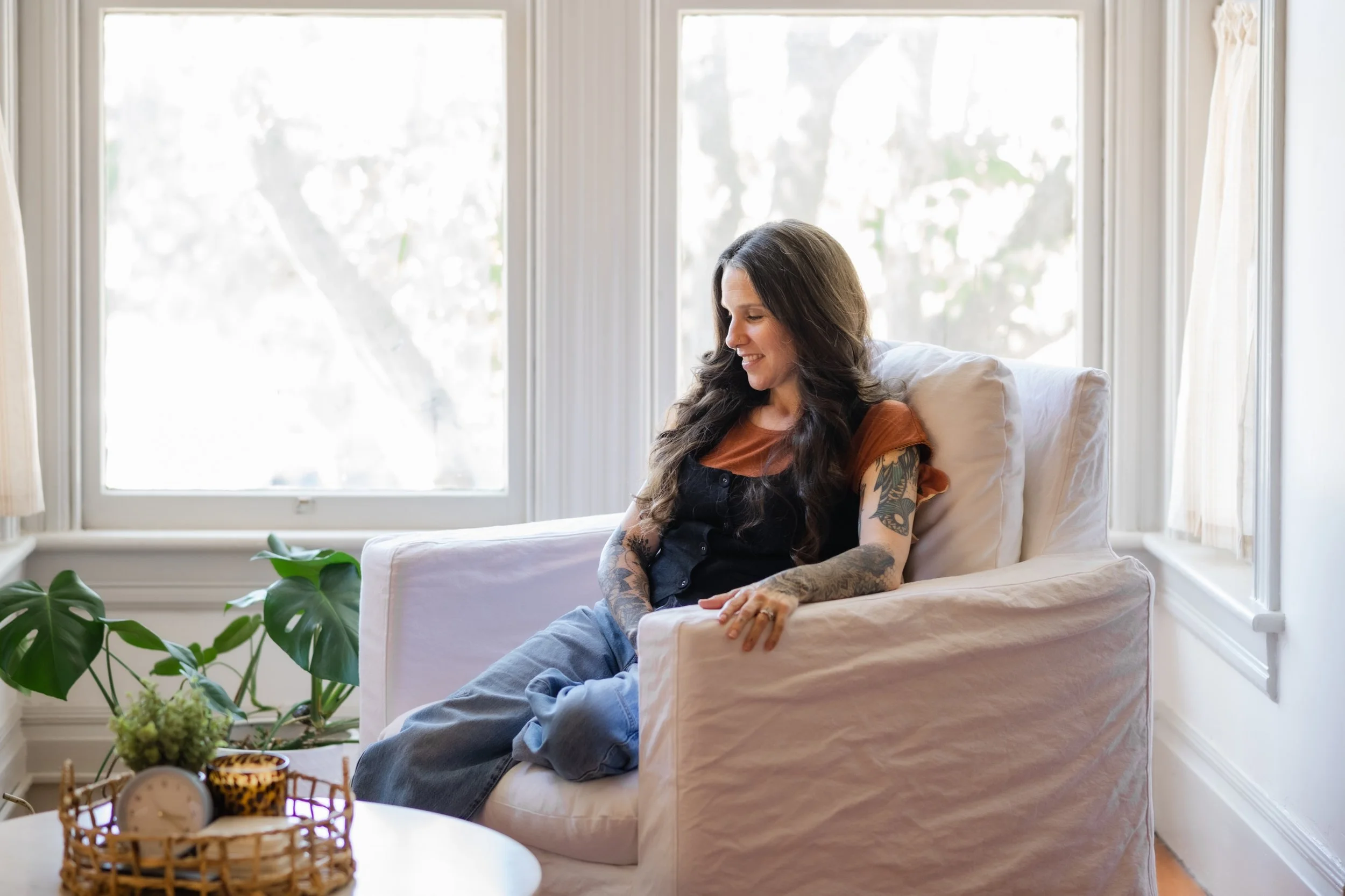 A woman with long dark hair and tattoos on her arms sitting on a white couch in front of a large window, smiling and looking down. There are plants and a basket on a table in the foreground.