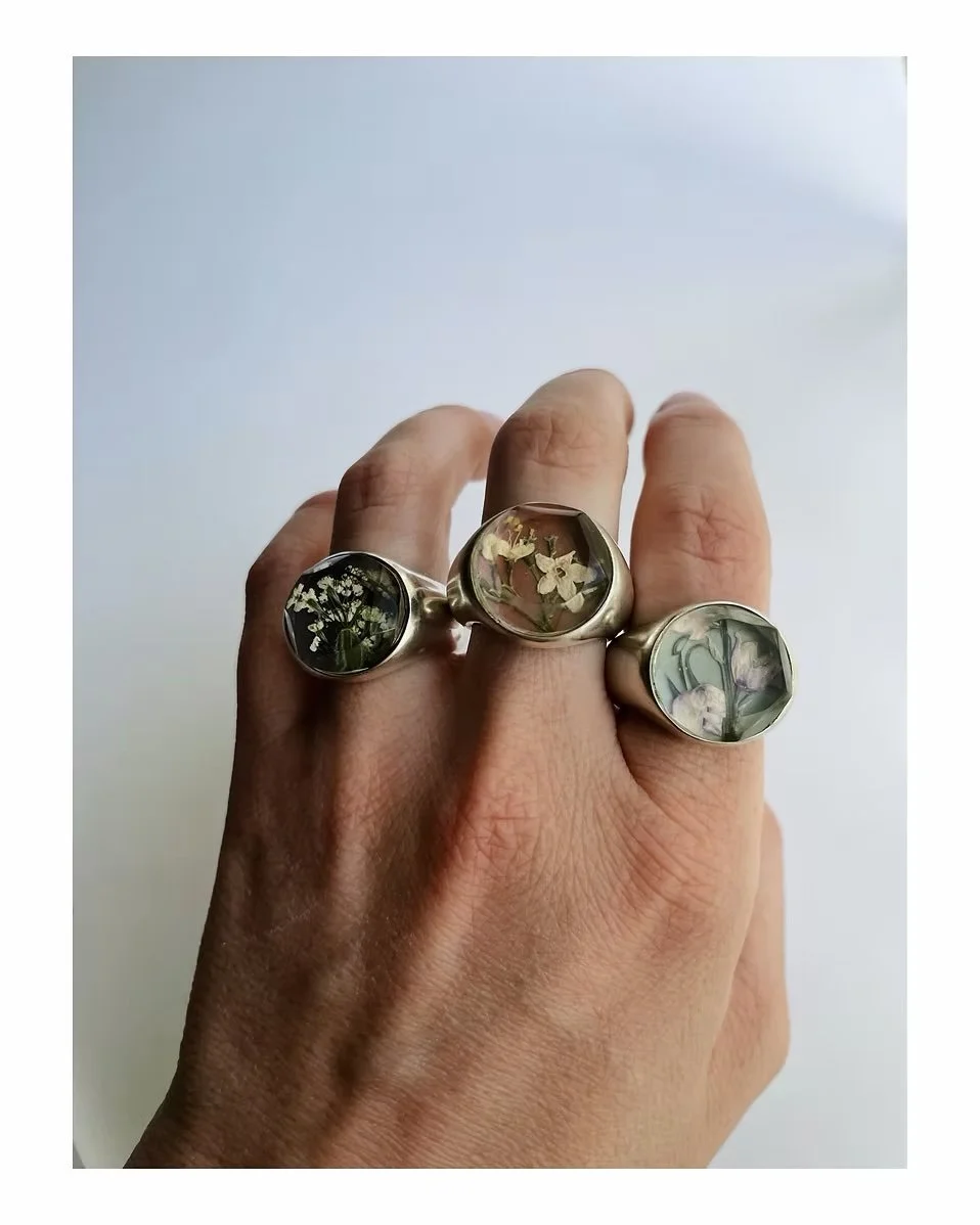 A hand wearing three silver rings with pressed flowers in glass cabochons, against a plain white background.