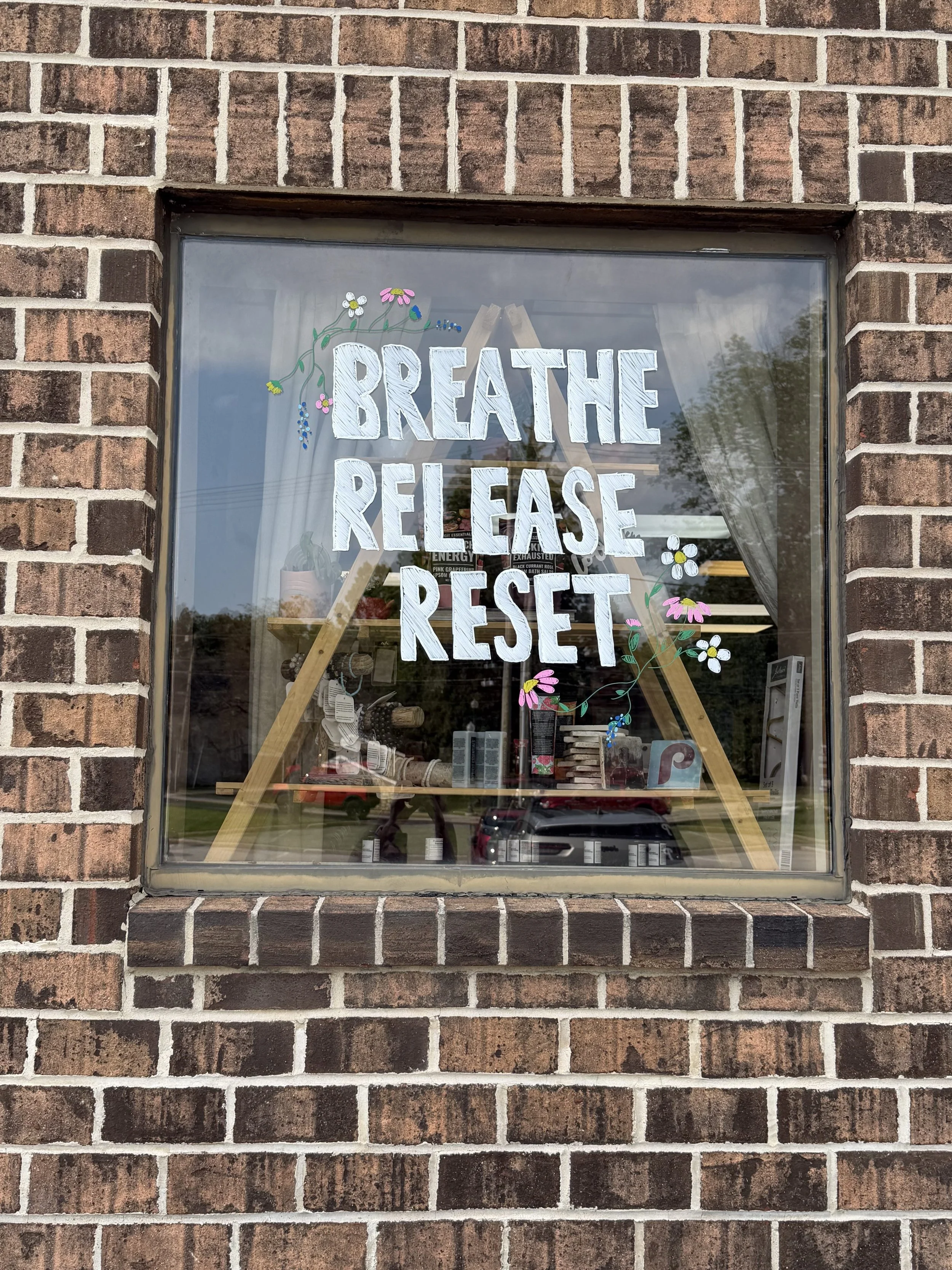A window on a brick building with a hand-painted sign that says, 'BREATHE RELEASE REST,' decorated with painted flowers and leaves.
