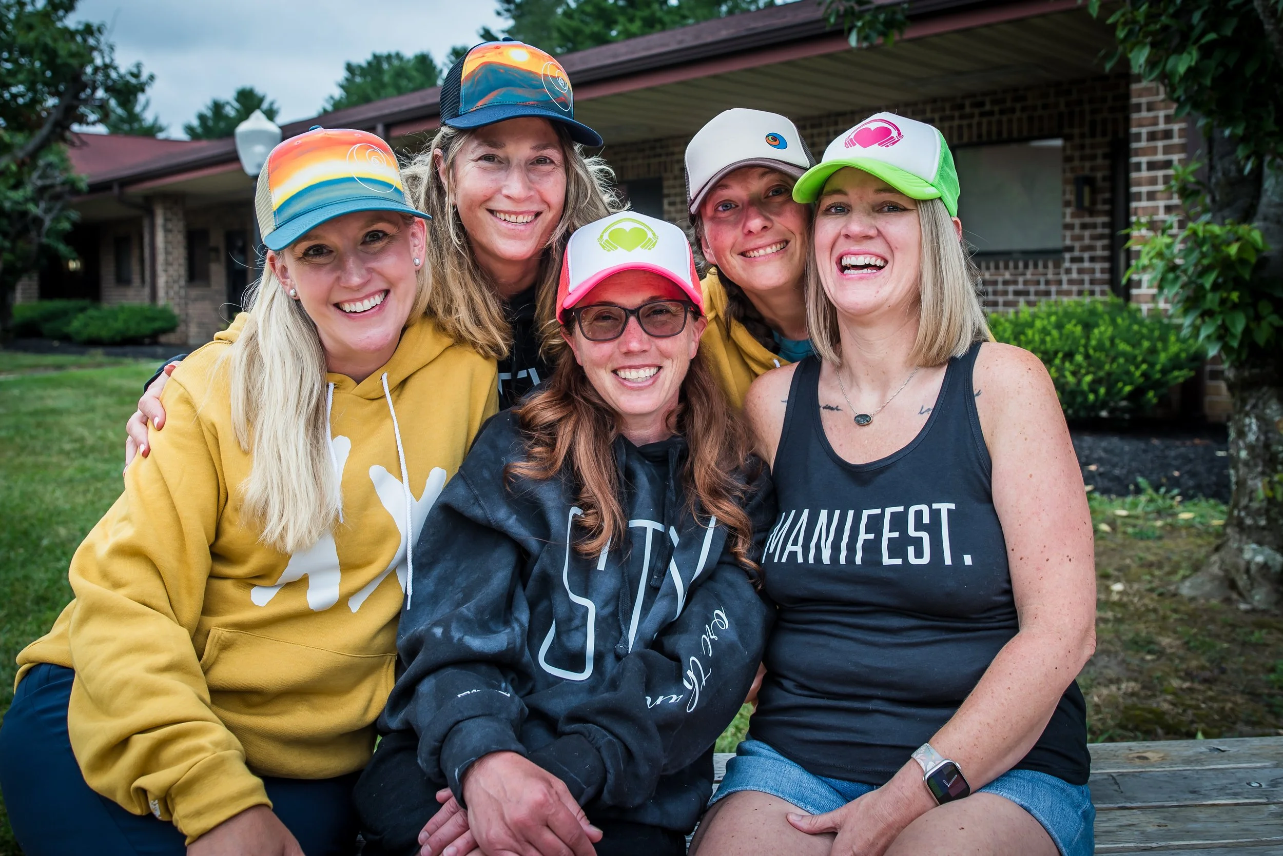 Six women smiling and posing together outdoors in front of a brick house and greenery, wearing casual clothing and hats.
