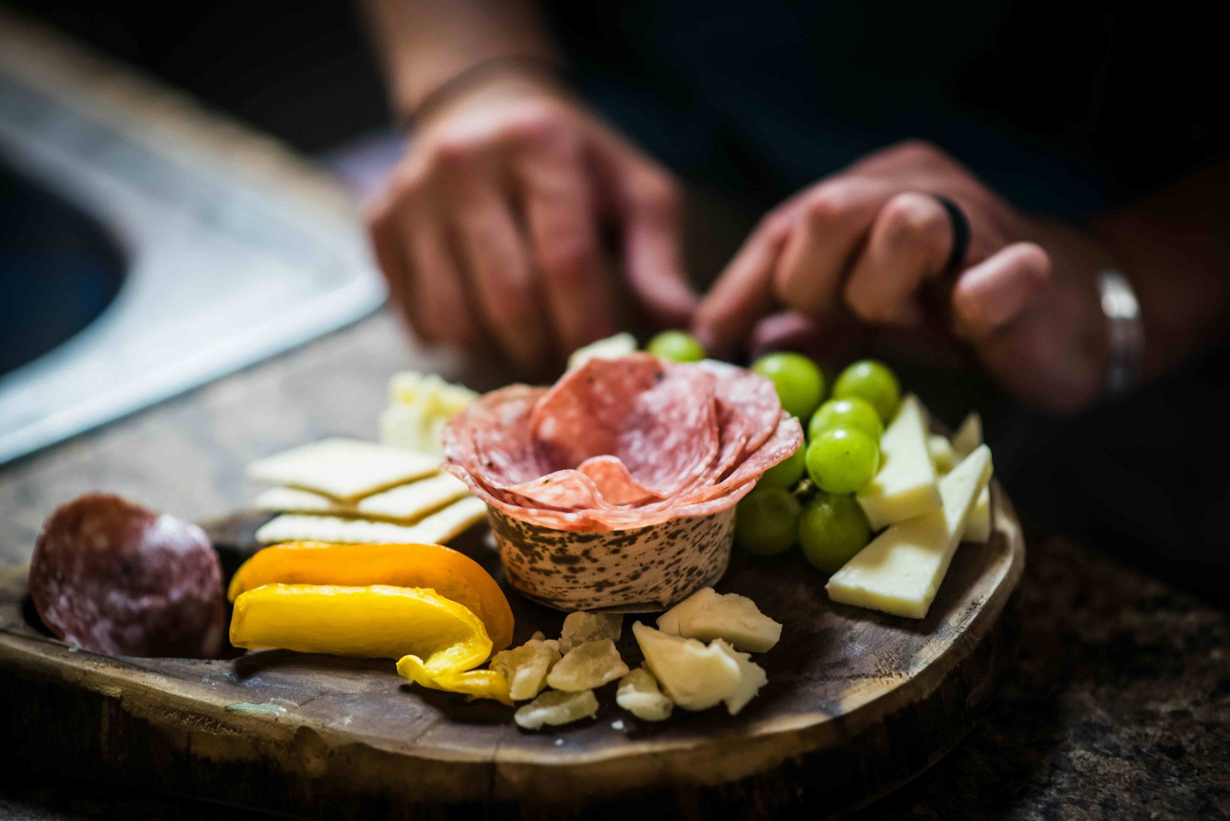 Close-up of assorted cheeses, cured meats, green grapes, and yellow bell pepper slices on a wooden serving platter, with hands and a blurred background.