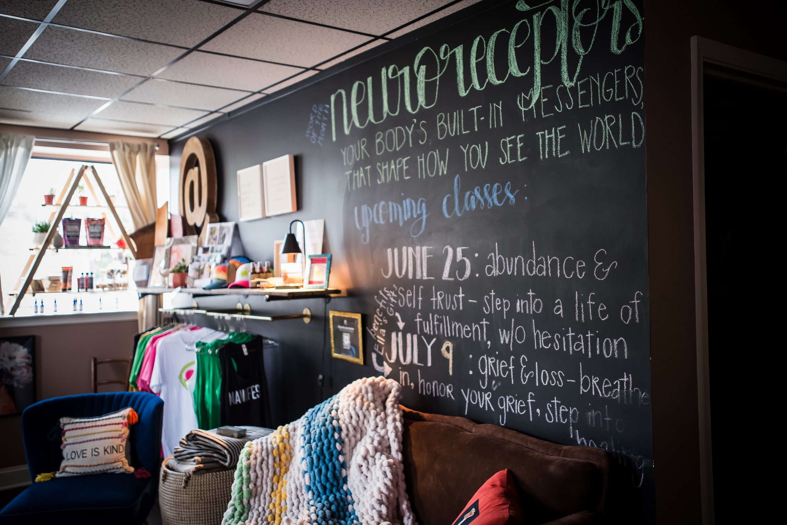Interior of a store or classroom with a chalkboard wall featuring white and colorful writing about upcoming classes and events. Shelves with potted plants, books, and products are visible in the background. A table displays various craft and knitting supplies, including yarn, pillows, and T-shirts.