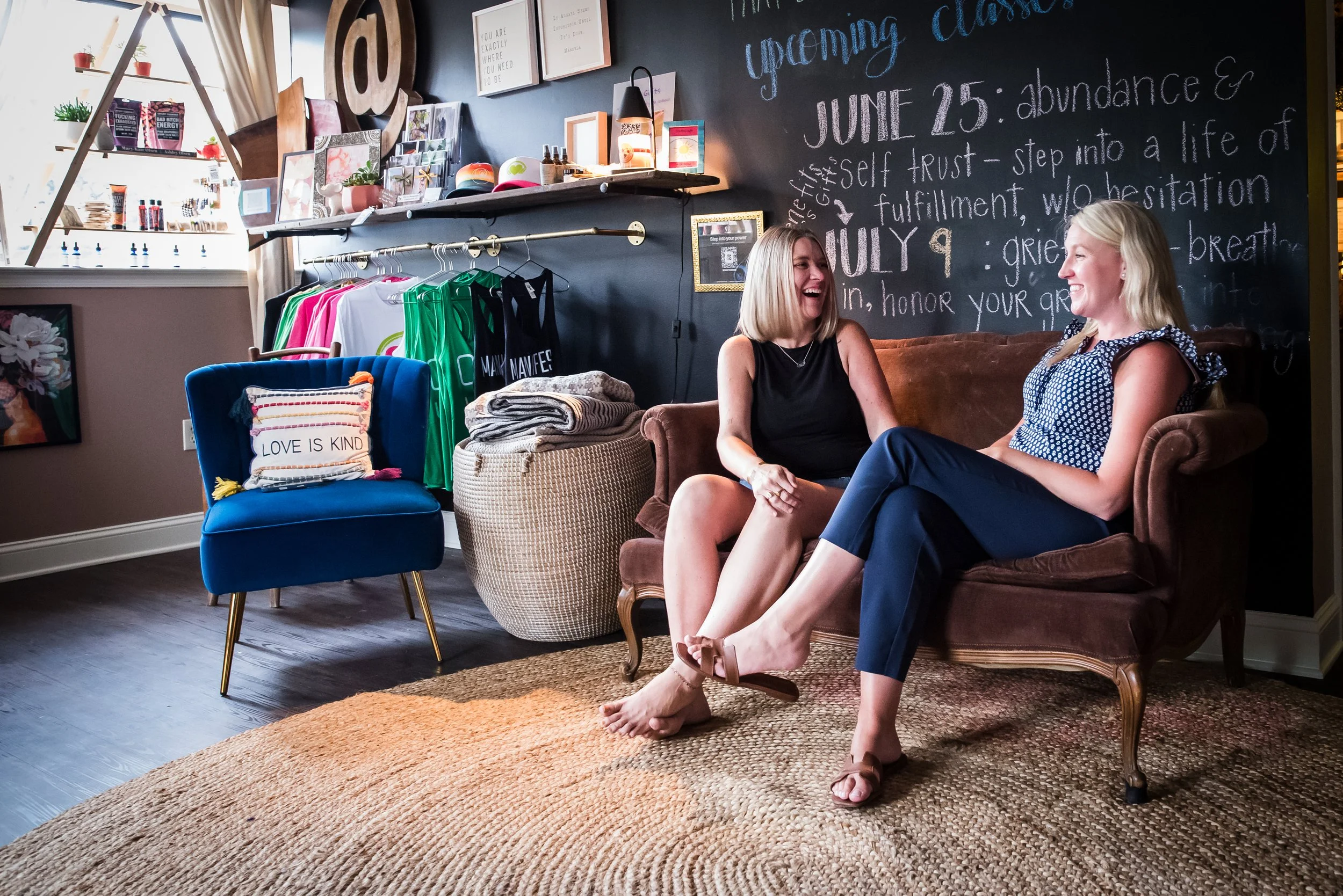 Two women sitting on a vintage brown couch, laughing and talking in a cozy, decorated room with a black chalkboard wall, a shelving unit with decorative items, and a clothing rack with colorful shirts.