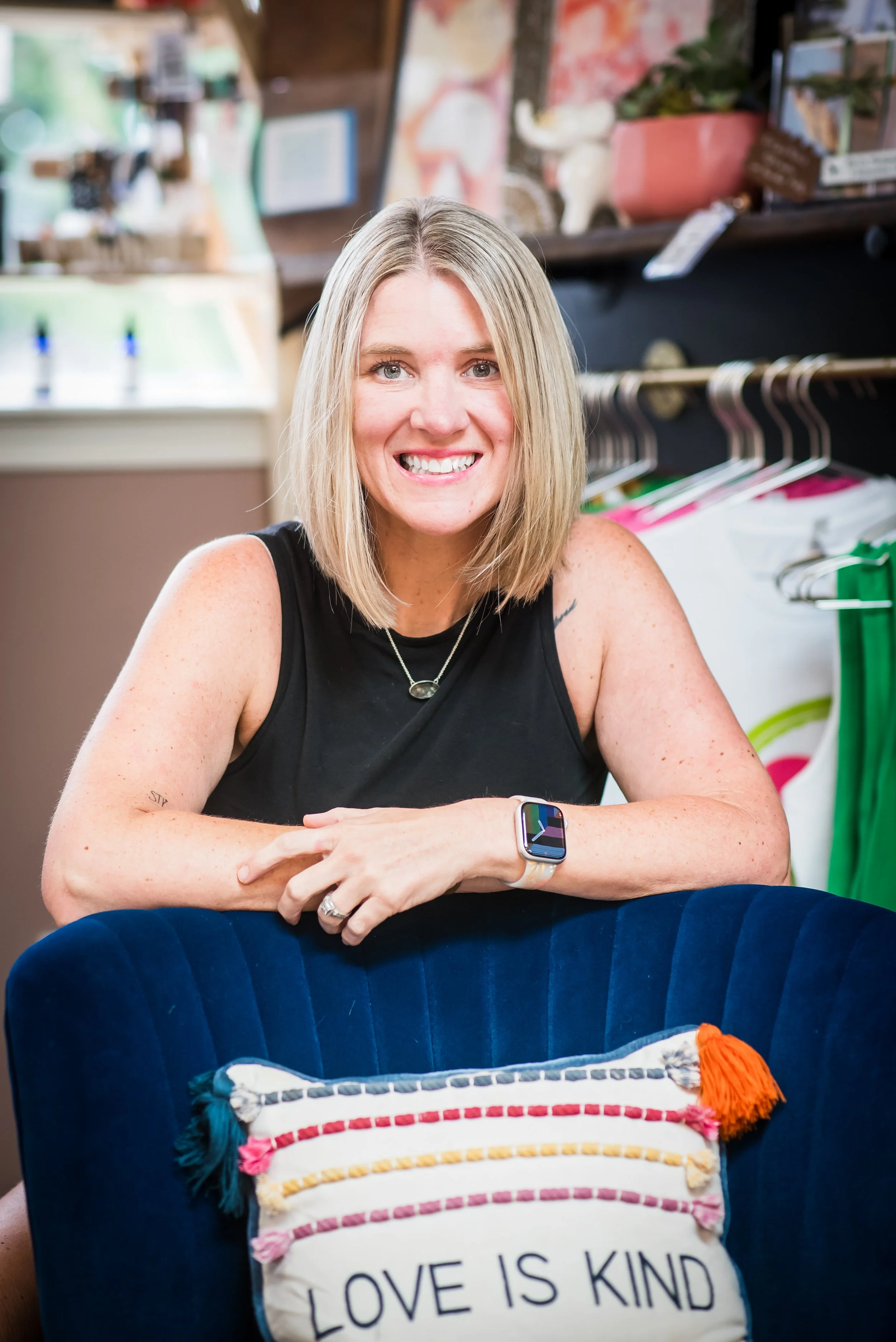 A woman with shoulder-length blonde hair, wearing a black sleeveless top, smiling at the camera, seated at a table in an indoor setting with colorful decor and clothing racks in the background.
