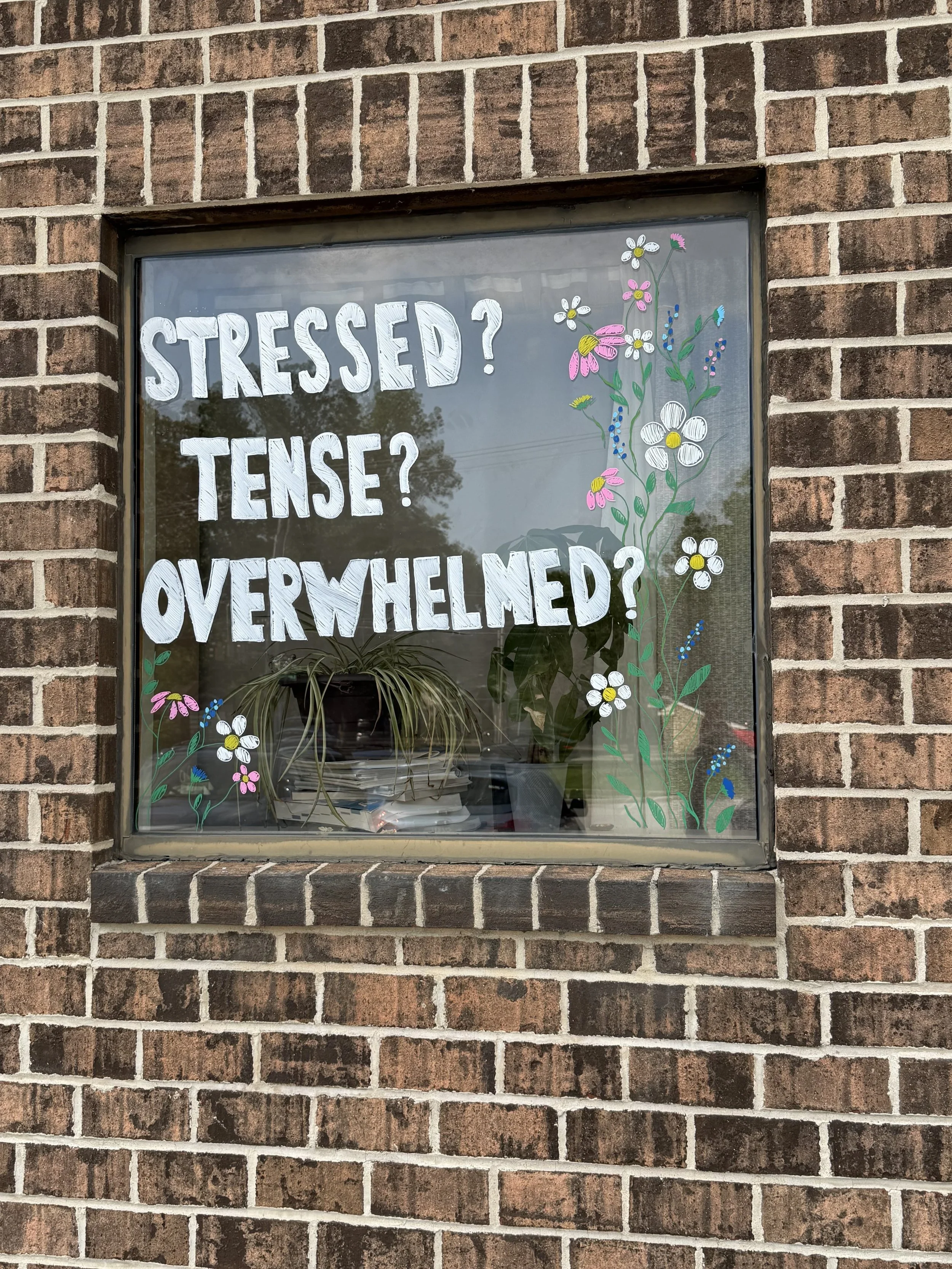 A brick wall with a window that has handwritten text and painted flowers on it. The text reads, 'Stressed? Tense? Overwhelmed?'