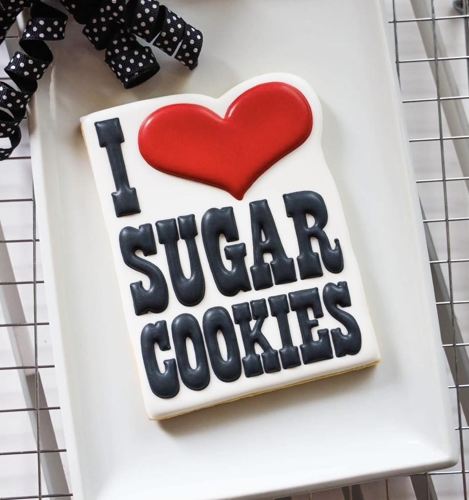 Decorative handmade cookie with the message "I love sugar cookies" and a red heart on top, placed on a white tray with a black ribbon with white polka dots.