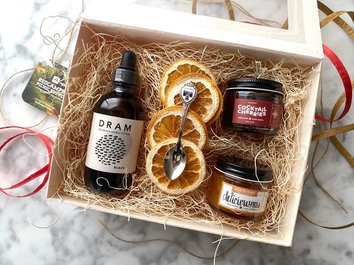 Gift box containing a black bottled tincture, dried orange slices with a small spoon, and two jars labeled 'Cocktail Cherries' and 'deliciousness,' surrounded by decorative ribbon.