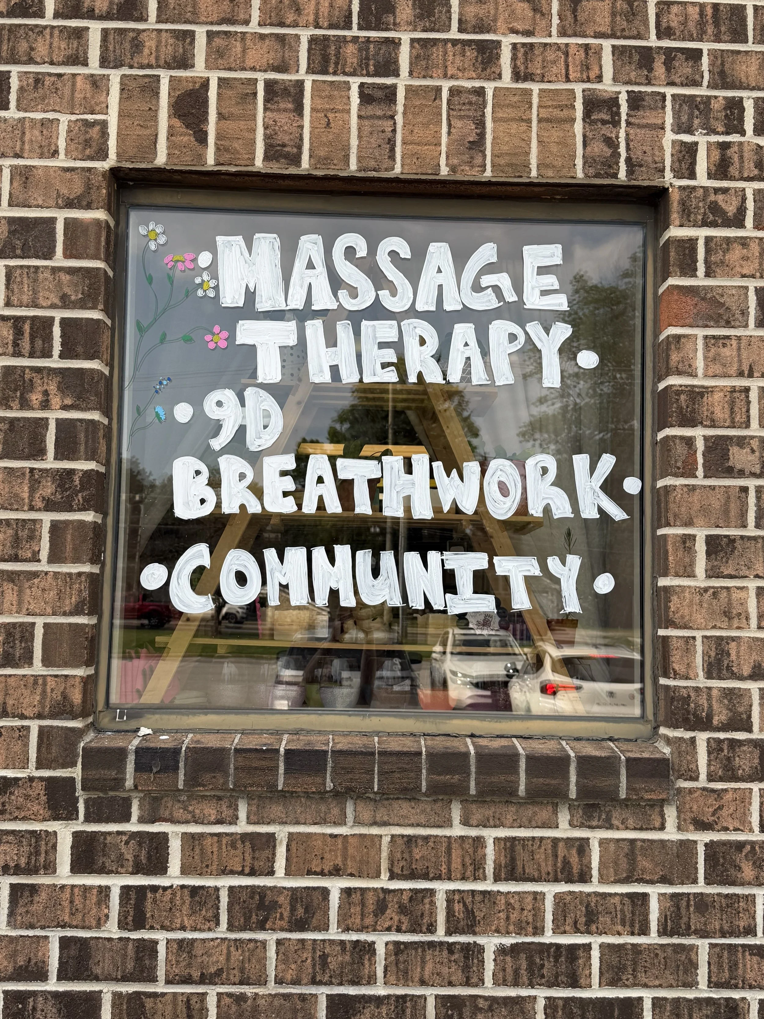 A window with hand-painted white letters advertising massage therapy, 9D breathwork, and community. The window is part of a brick building, and there are small decorative flowers painted near the text.