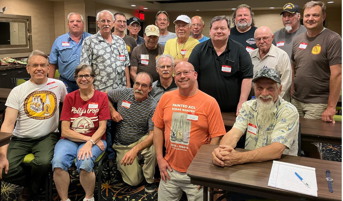 Group of 16 adults gathered in a room, posing for a photo, with some sitting and others standing behind them, smiling and wearing casual clothes with name tags.