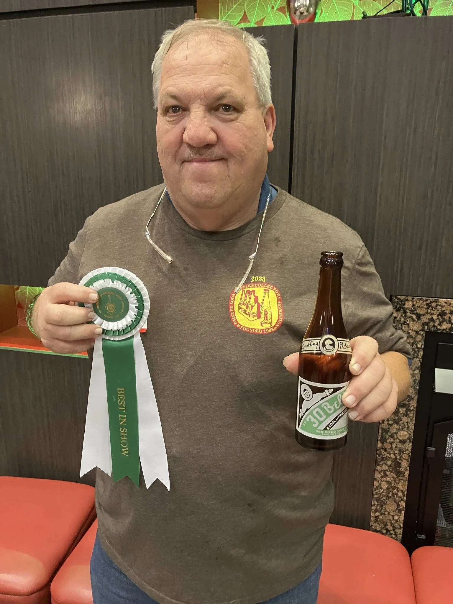 A man with gray hair holding a beer and a green and white ribbon award. He is wearing a gray shirt with a red and yellow logo on the chest. The background includes a dark wood partition and red seating.