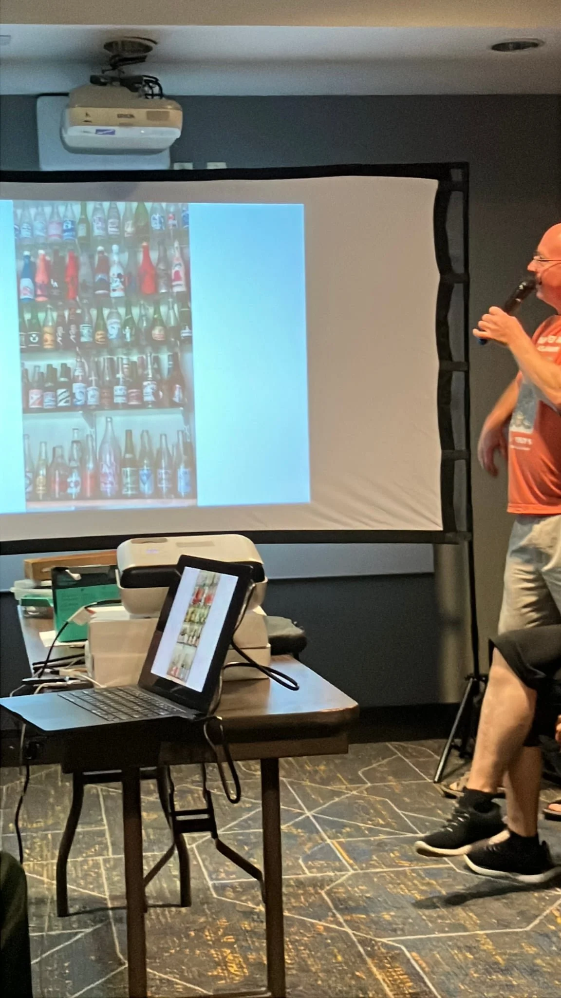A man standing and speaking into a microphone during a presentation, with a projection screen displaying an image of various bottles behind him. The room has laptops on a table, and the floor features a dark, patterned tile.