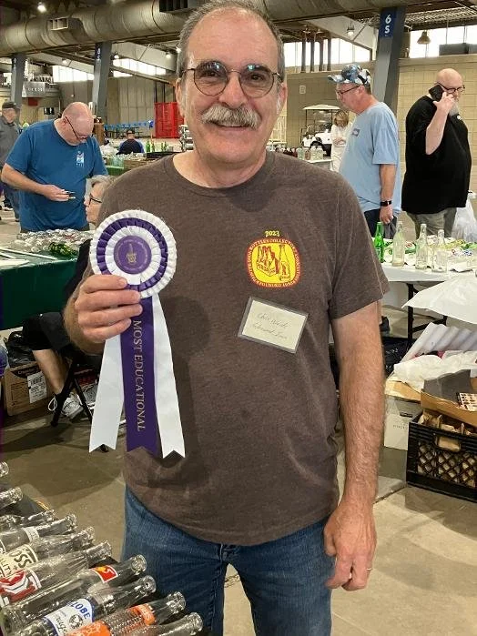 A man with glasses and a mustache is smiling and holding a purple ribbon award. He is wearing a gray T-shirt with a yellow patch and a name tag. The setting appears to be an indoor event or expo with tables, bottles, and other people in the background.