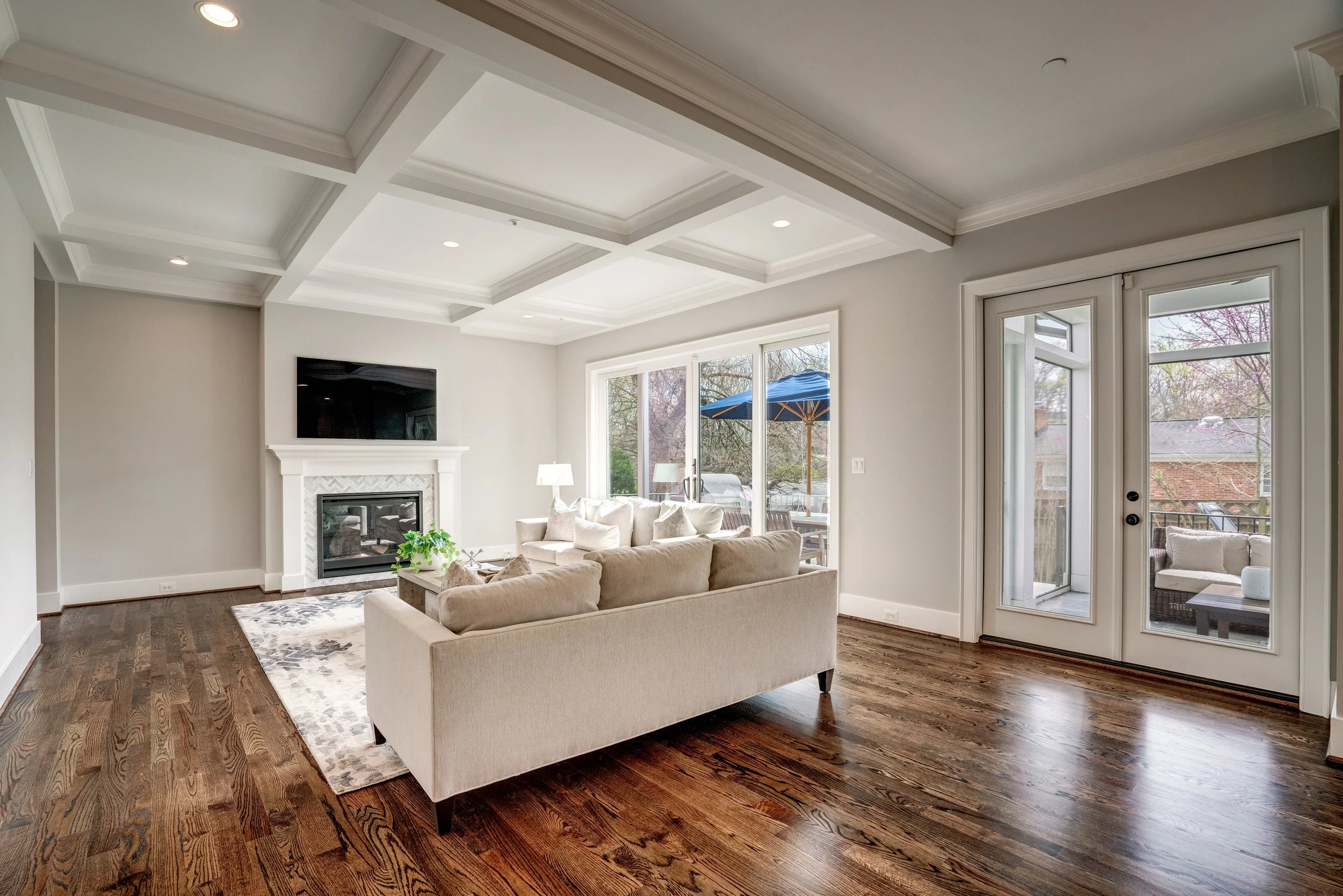 Bright living room with white walls, coffered ceiling, wooden floors, a beige sofa, a fireplace, large windows, and a sliding glass door leading to an outdoor patio.