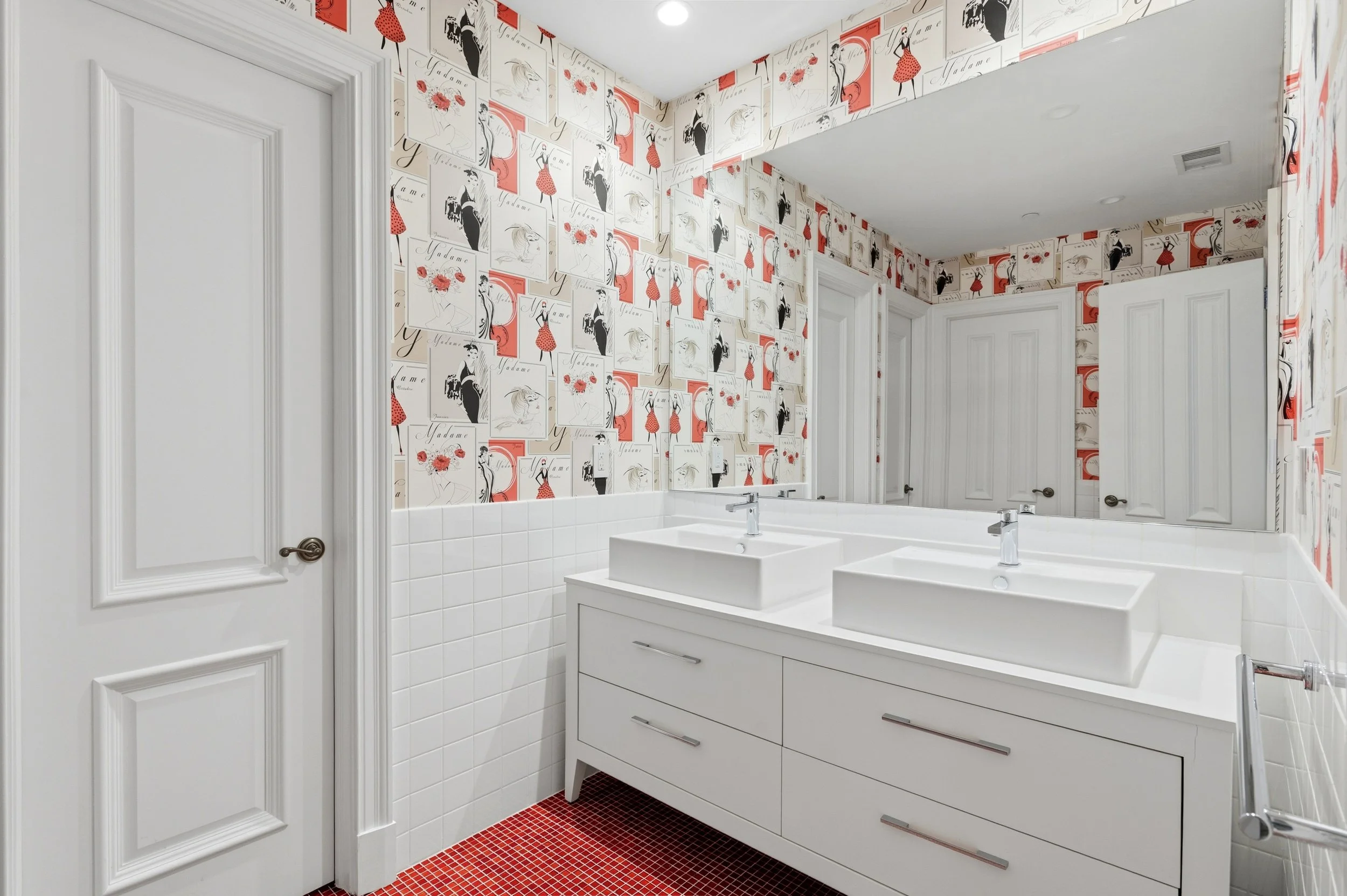 A modern bathroom with a twin sink vanity, white cabinets, and a large mirror. The walls are decorated with patterned wallpaper featuring illustrated women in red dresses and black outfits, with the word 'Madame' written. The floor has red mosaic til