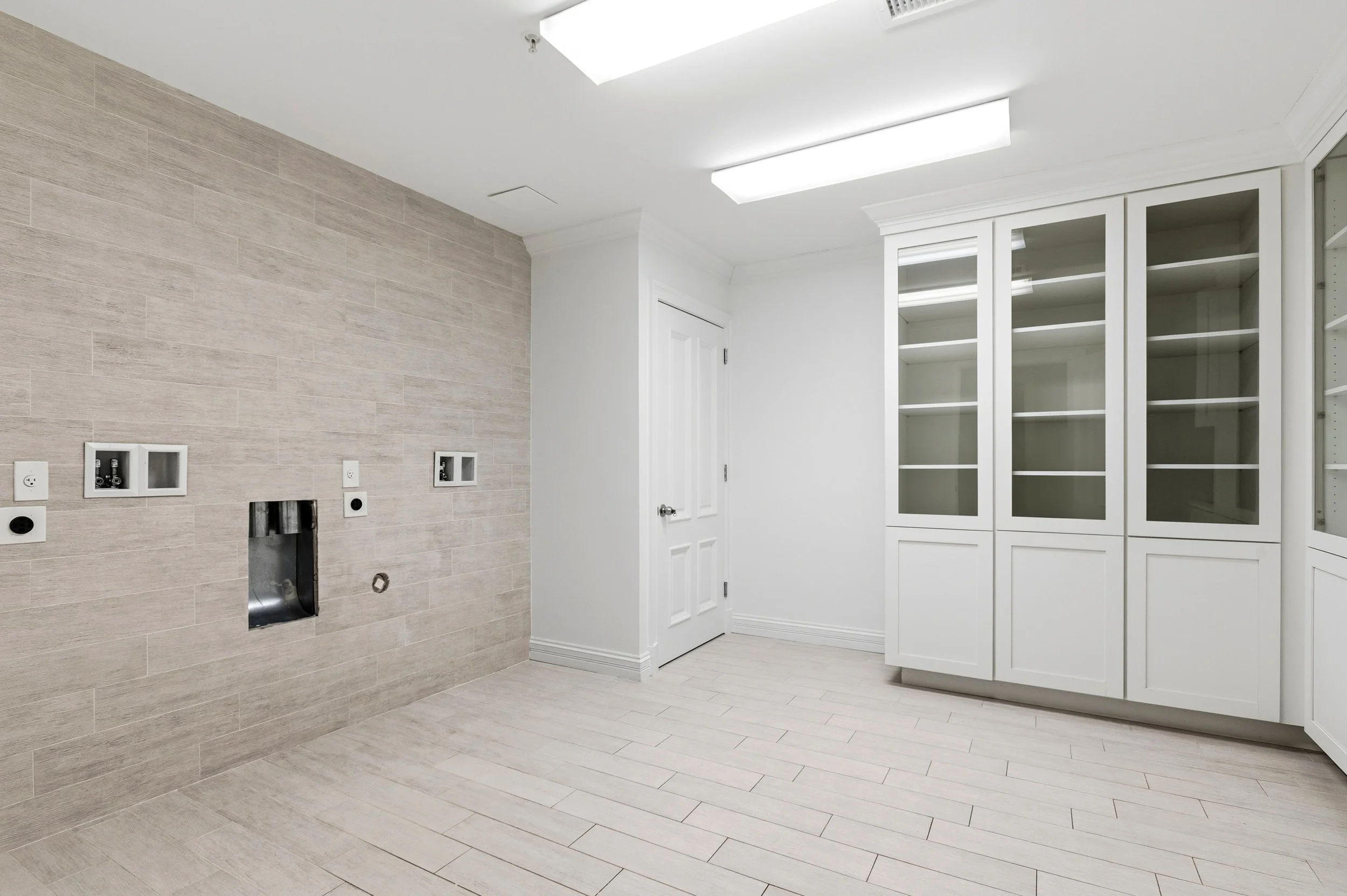 Empty laundry room with beige tiled floors and walls, white cabinetry with glass doors, ceiling lights, and wall outlets.