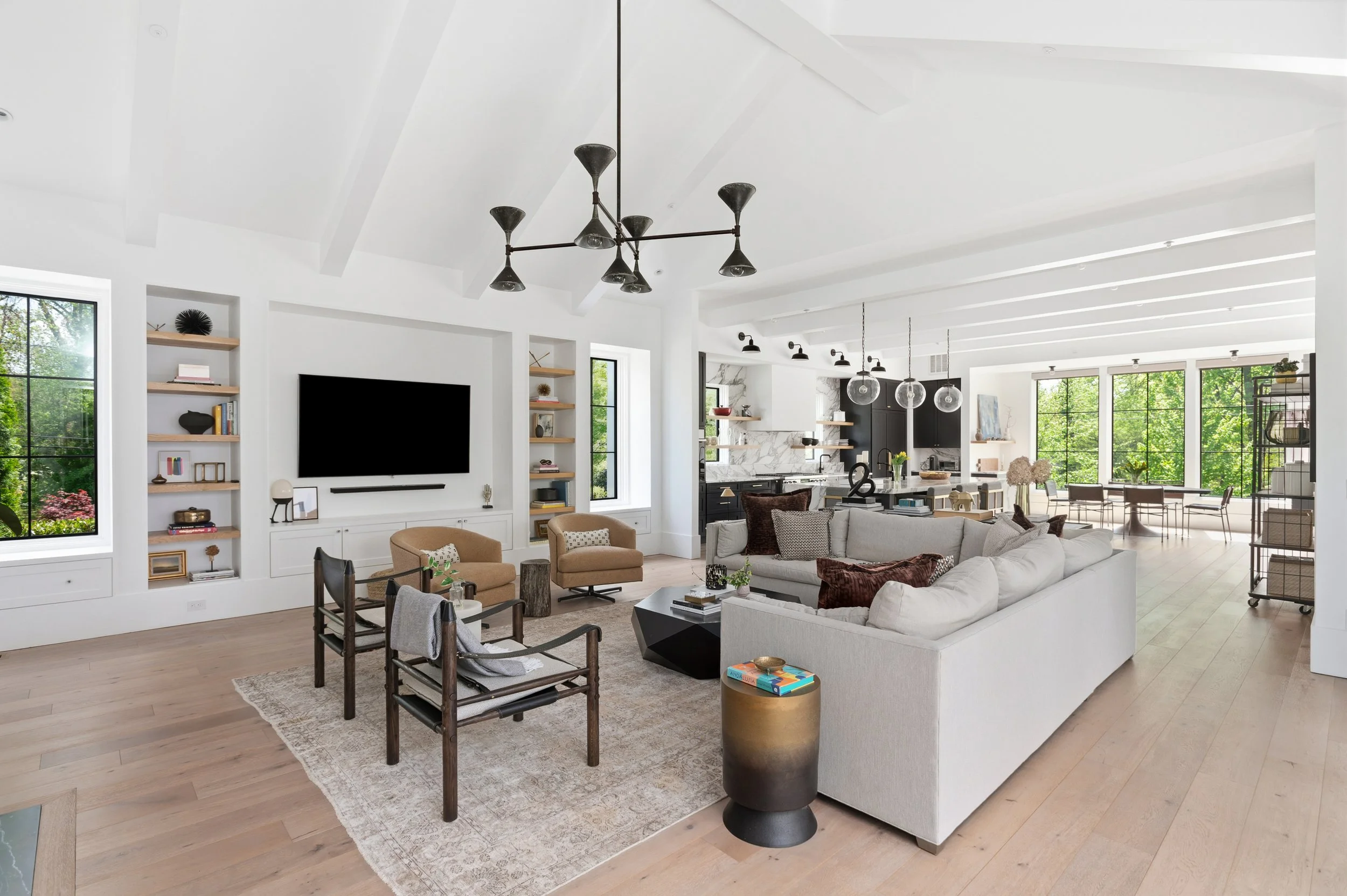 A spacious, modern living room with white walls and high ceilings featuring exposed beams, large windows with black frames, and a mix of contemporary and rustic furniture, including a large white sectional sofa, wooden chairs, and a black and gold si