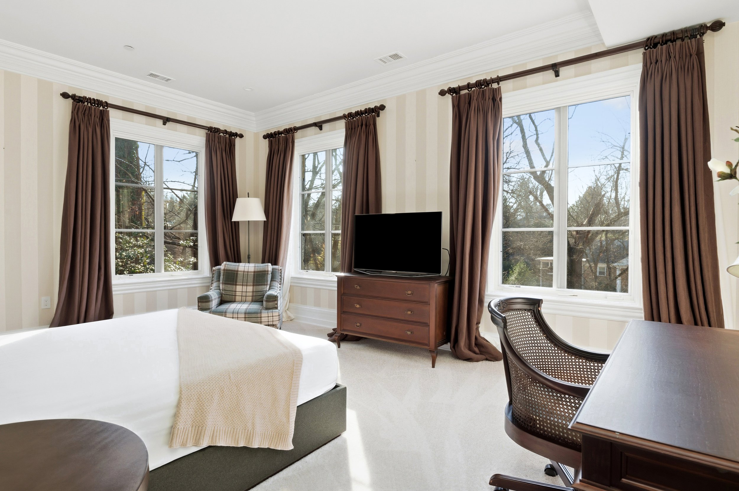 Living room with three large windows, brown curtains, a plaid armchair, a flat-screen TV on a wooden dresser, a white sofa with a beige throw blanket, a dark round table, and a curved wooden desk with a woven chair. Outside the windows are trees and 