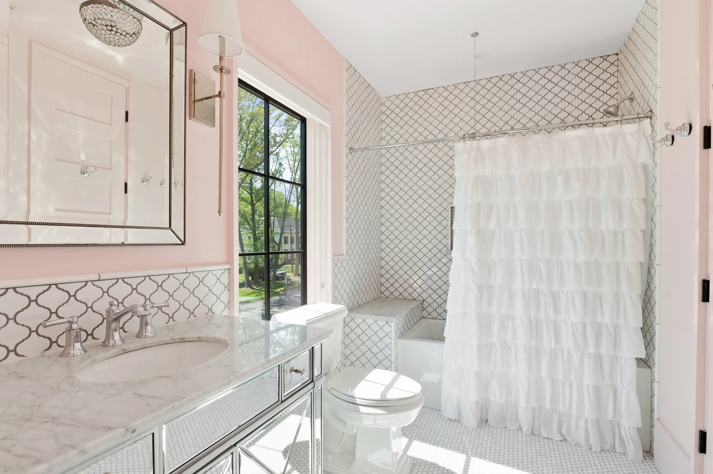 A bright bathroom with a large window showing green trees outside. The walls are pink, accented with black and white patterned tiles. The bathroom features a white vanity with a marble countertop, a mirror with a beaded border, and a white shower cur