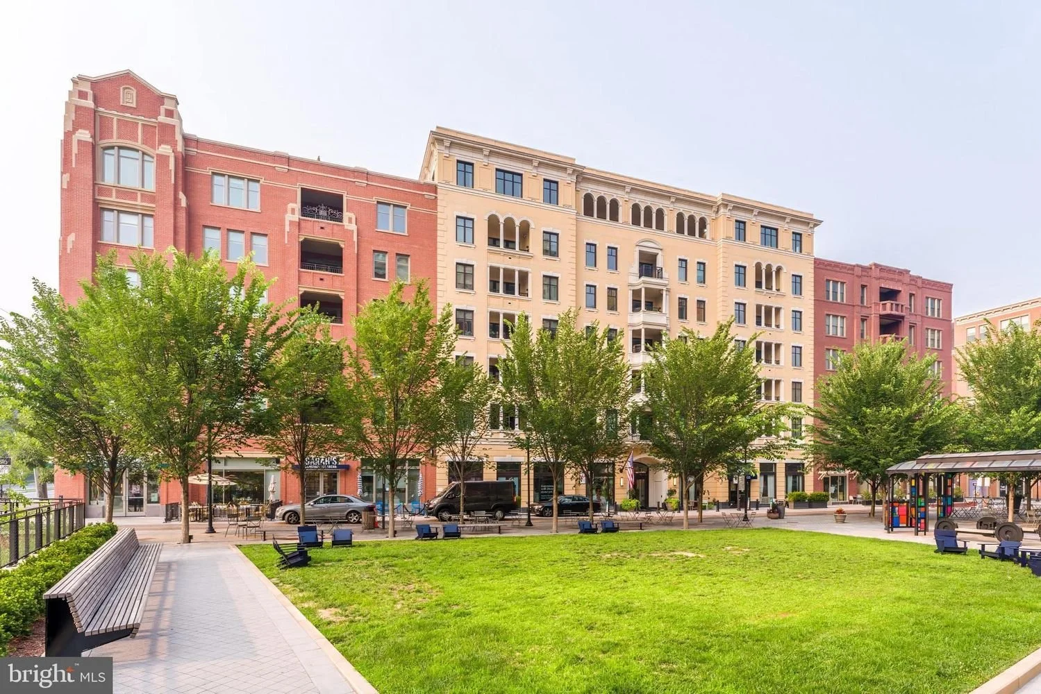 A park with a grassy area, benches, trees, and a walking path, in front of a multi-story residential building with shops on the ground floor.