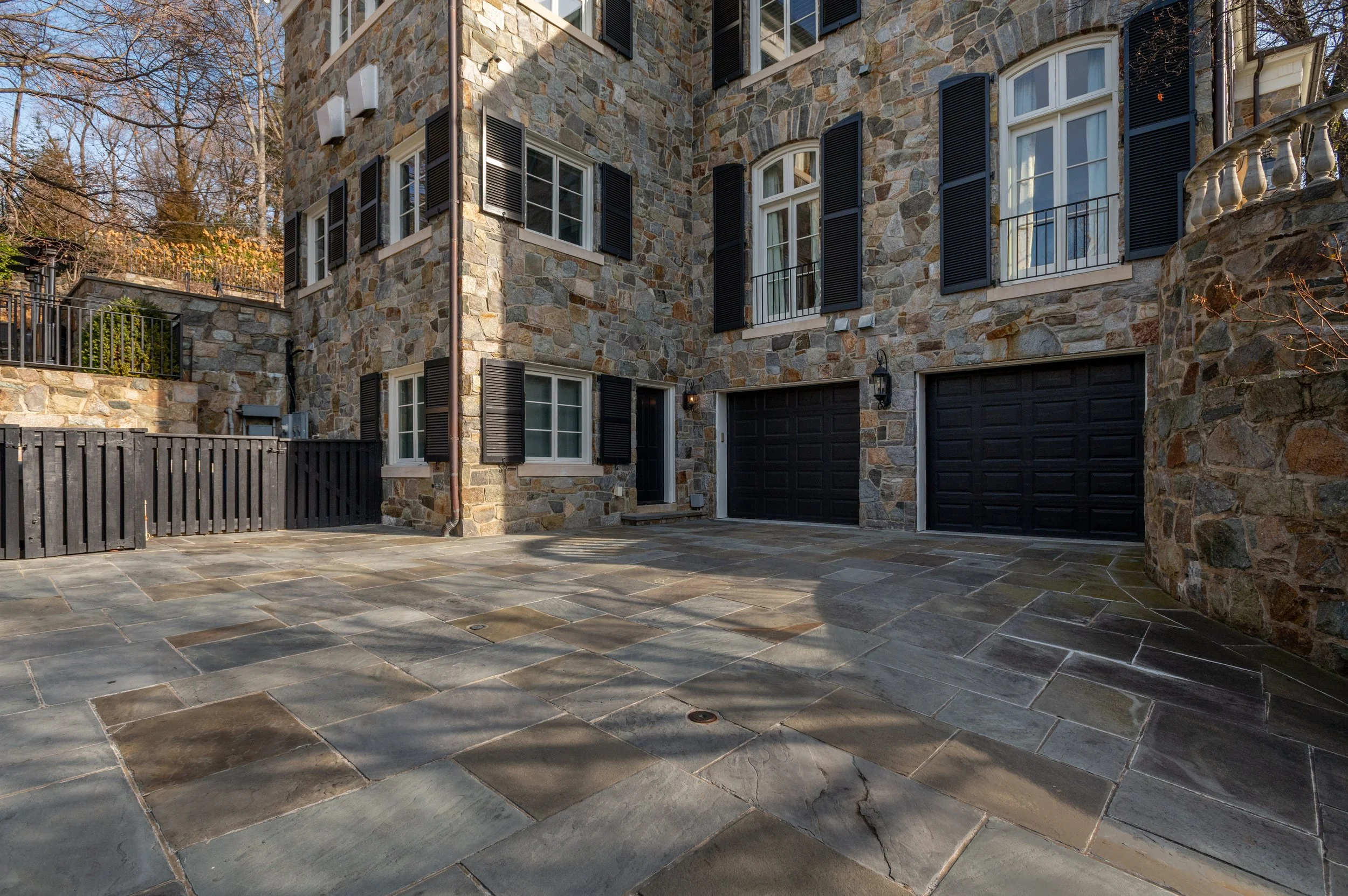A stone building with black shutters and white window frames, black garage doors, and a tiled patio outside, with bare trees in the background.
