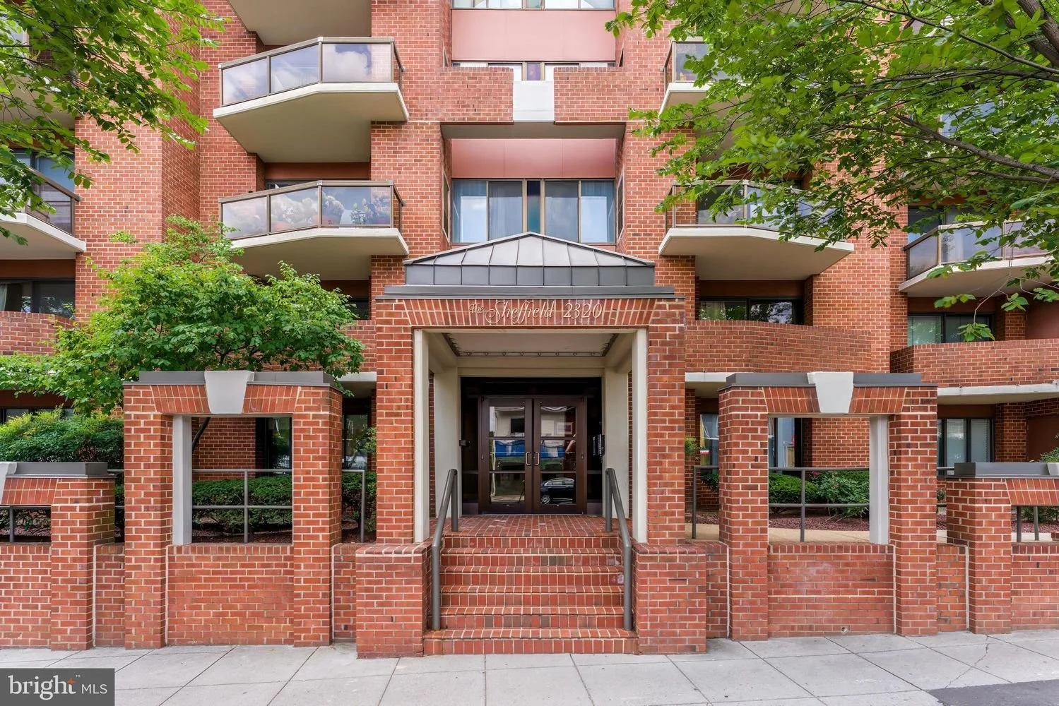 Red brick apartment building with small balconies, trees, and a gated entrance with stairs.