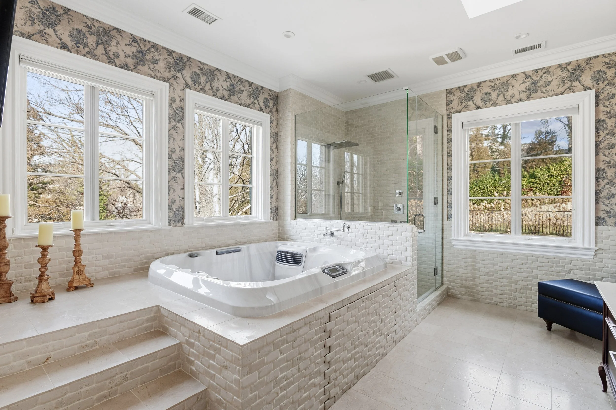Bathroom with large window, a corner shower with glass door, and a whirlpool tub. Decor includes candles on candlesticks and a blue upholstered stool.