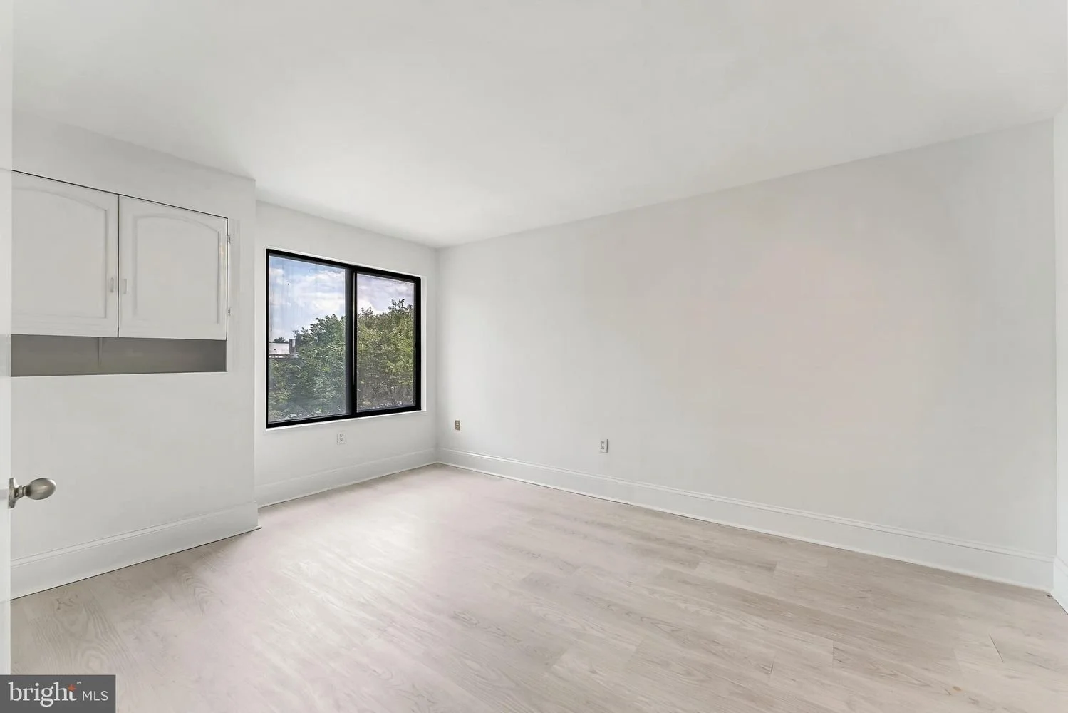 Empty room with white walls, light-colored wooden floor, black-framed window showing trees outside, and white cabinets on the left wall.