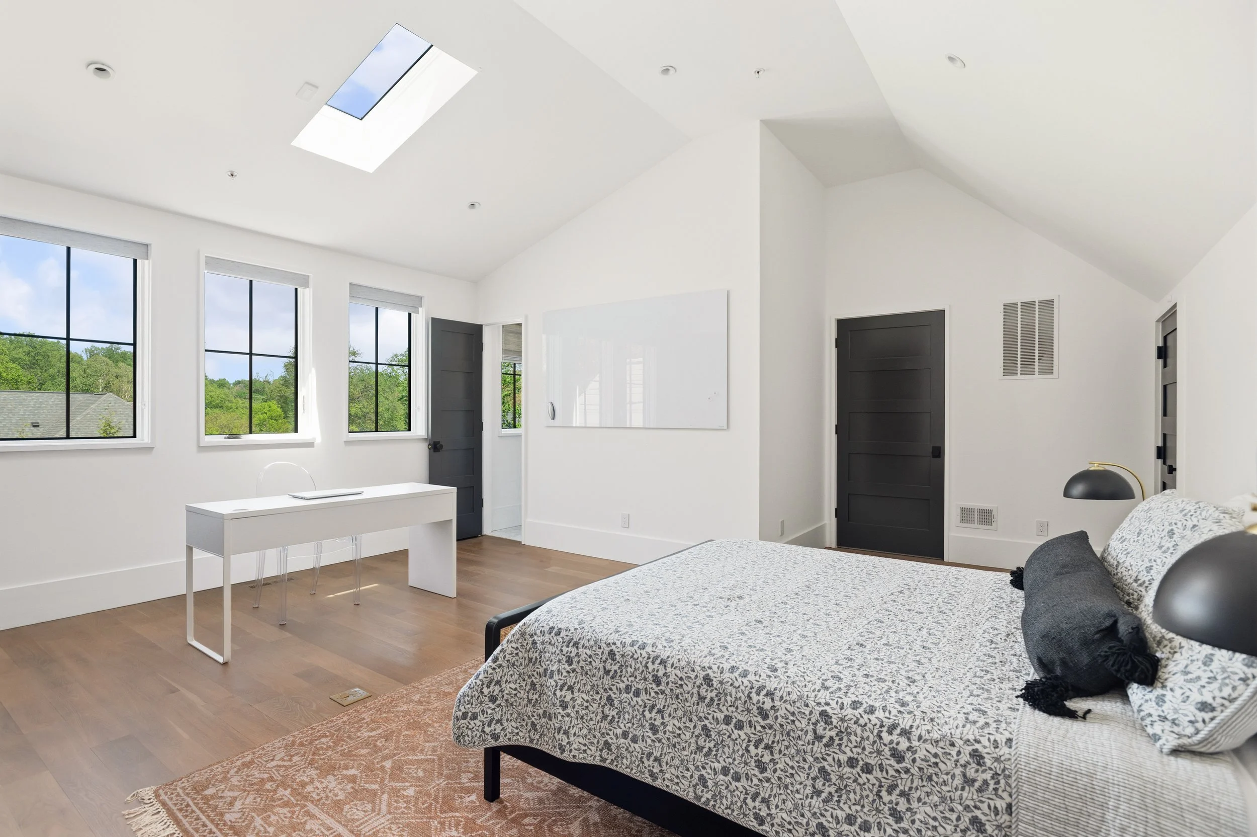 Modern bedroom with white walls, large windows, and a skylight in the ceiling. Contains a bed with a patterned bedspread, a white desk with a clear chair, and dark doors with window accents. Hardwood floor and an orange rug under the bed.