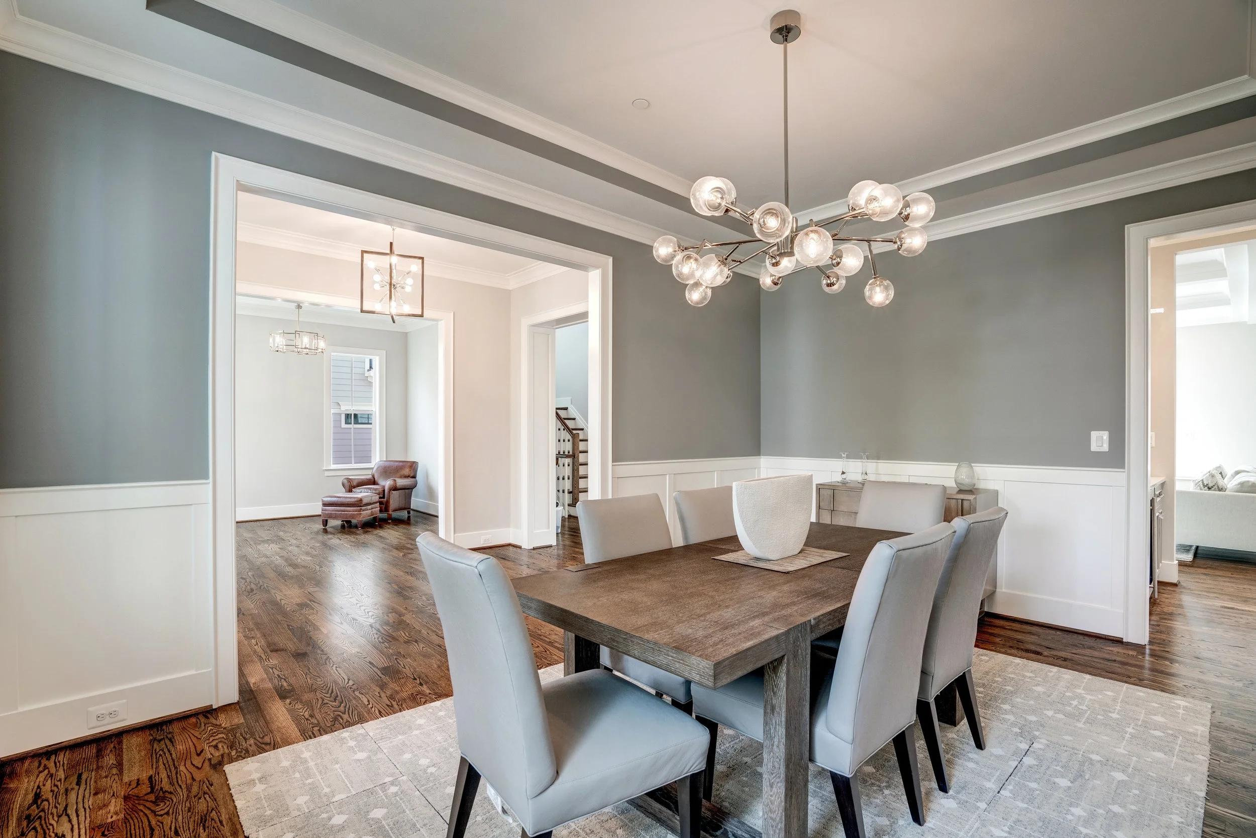 A dining room with a wooden table, light gray upholstered chairs, a modern chandelier with multiple glass globes, and hardwood floors. There's a view into other rooms with additional lighting fixtures and windows.