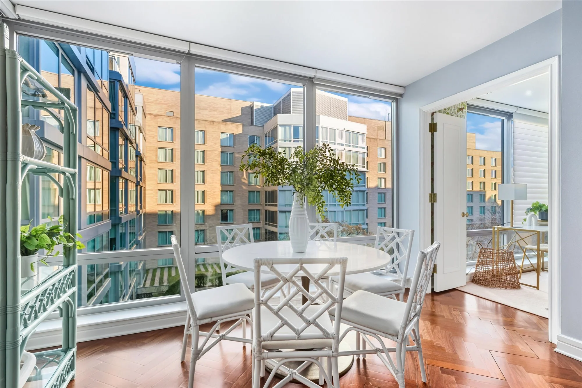 Bright sunlit room with large windows, white round dining table with white chairs, potted plants, and a view of neighboring apartment buildings.
