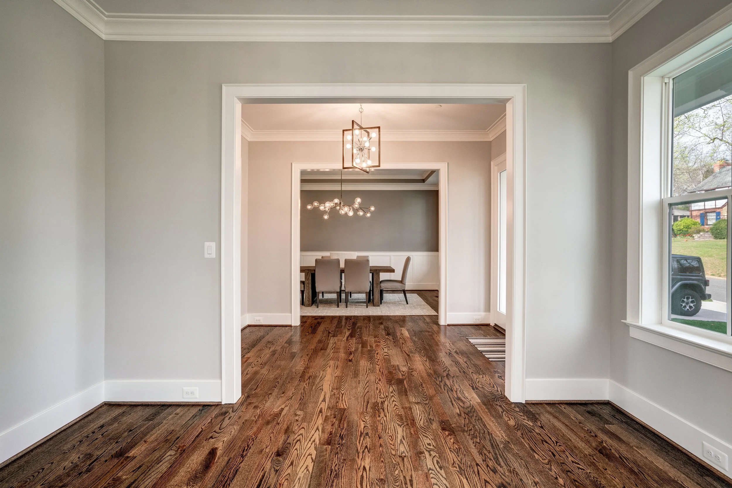 Interior view of a house with hardwood flooring, white walls, doorway leading to a dining room with a light fixture, window on the right showing a car and outside greenery.