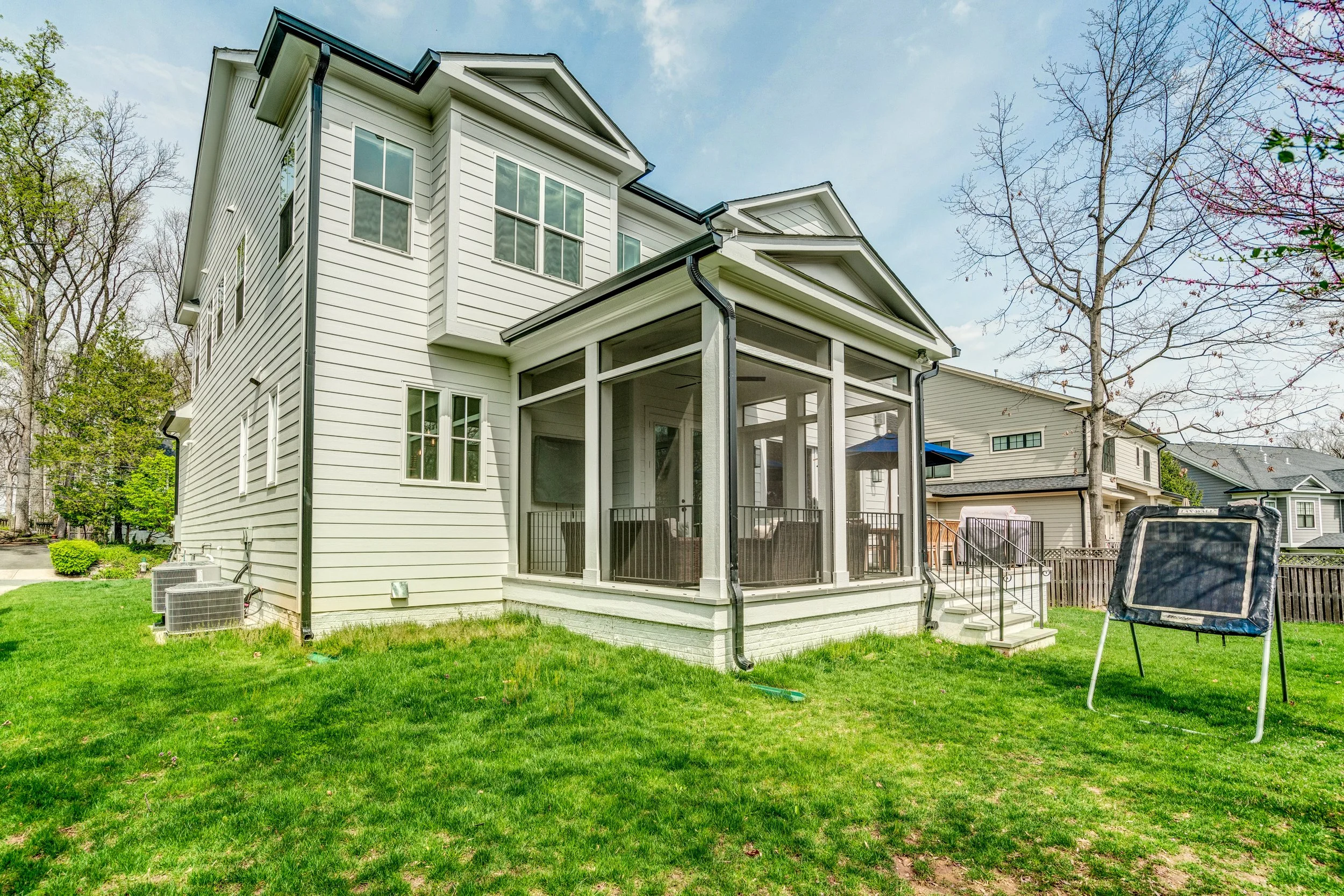 Backyard view of a multi-story house with a screened porch, surrounded by a green lawn, trees, and neighboring houses.