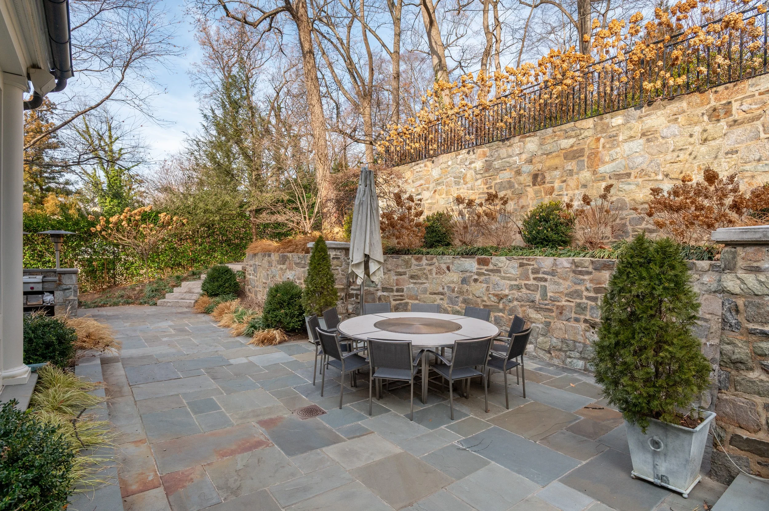 An outdoor patio area with stone flooring, a round dining table with chairs, and a stone wall with plants and trees in the background, during late autumn.