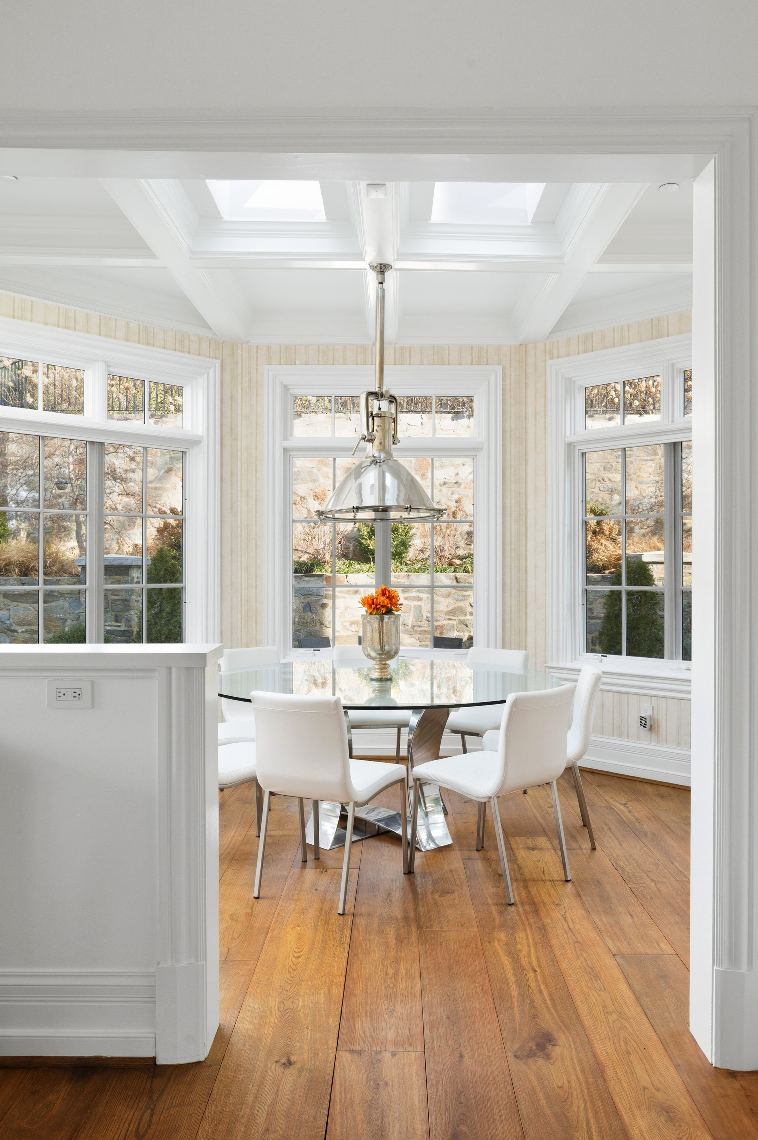 A dining room with a round glass table, white chairs, a metal pendant light, large windows, and hardwood flooring, with a sunny outdoor view.