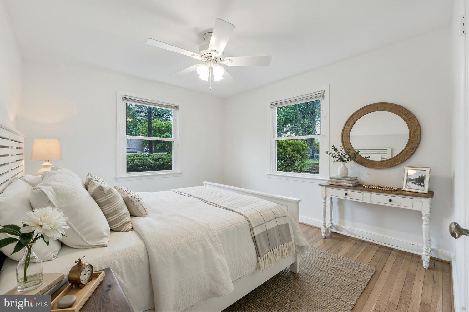 A bright bedroom with white walls, a white bed with multiple pillows, a side table with a flower, clock, and books, two windows showing greenery outside, a white ceiling fan, and a white and wood console table with decorative items and a large round 