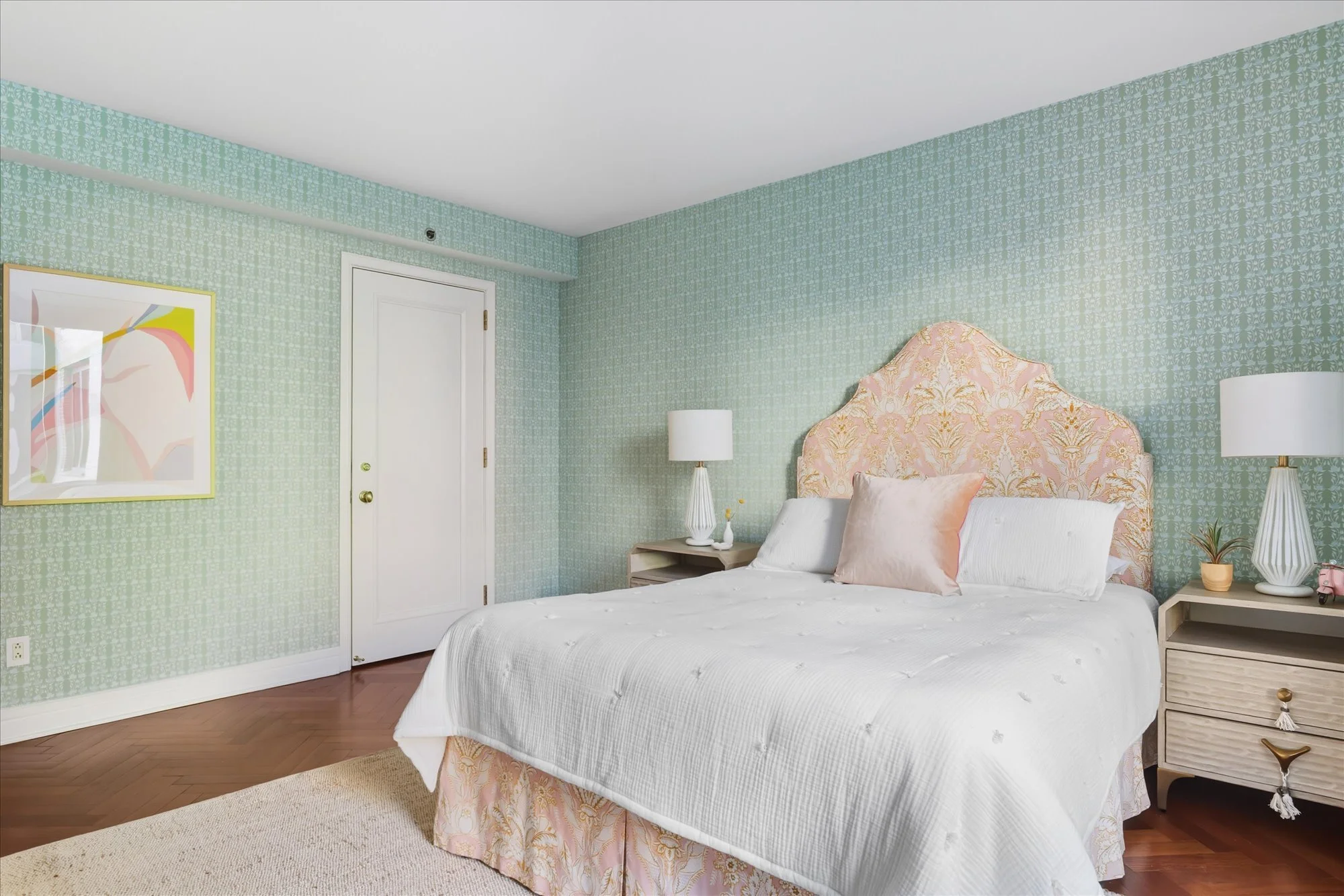 A bedroom with light green floral patterned wallpaper, a white door, a bed with white bedding, pink floral headboard, and matching bed skirt, two white lamps on nightstands, and a framed abstract art piece on the wall.